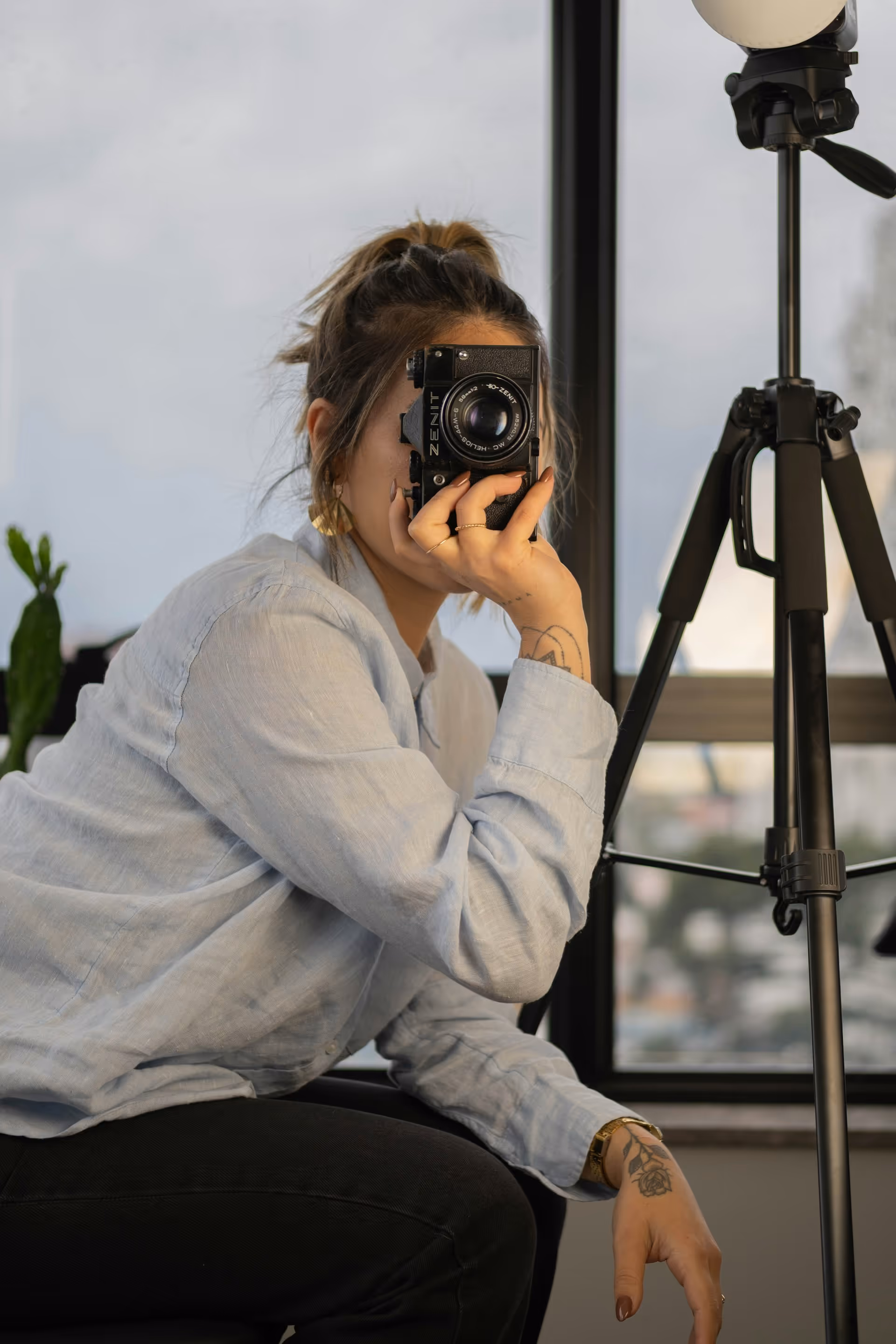Woman with tied-back hair holding a vintage camera in front of her face, sitting next to a tripod indoors.