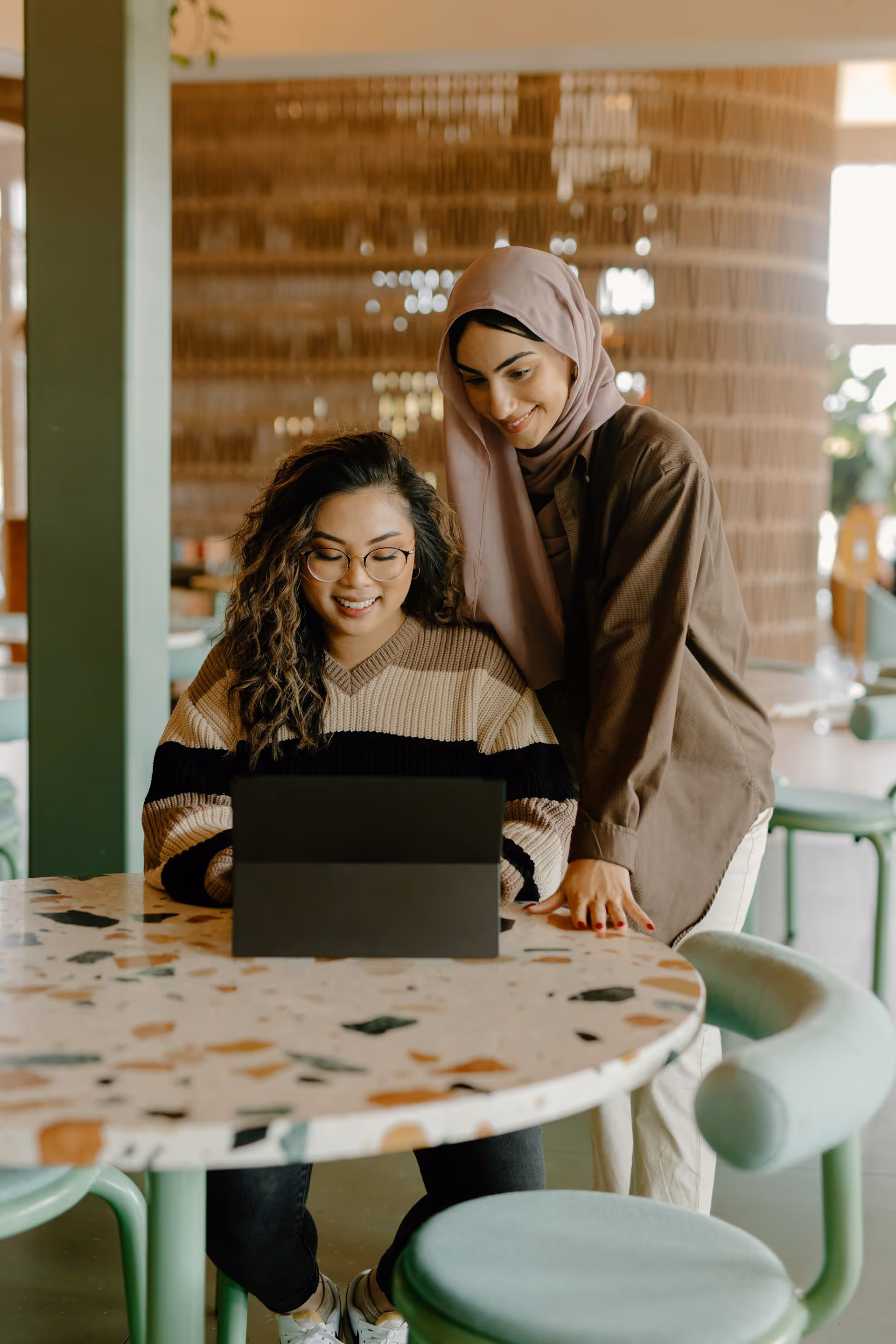 Two women smiling and looking at a tablet while sitting at a terrazzo table in a casual indoor setting.