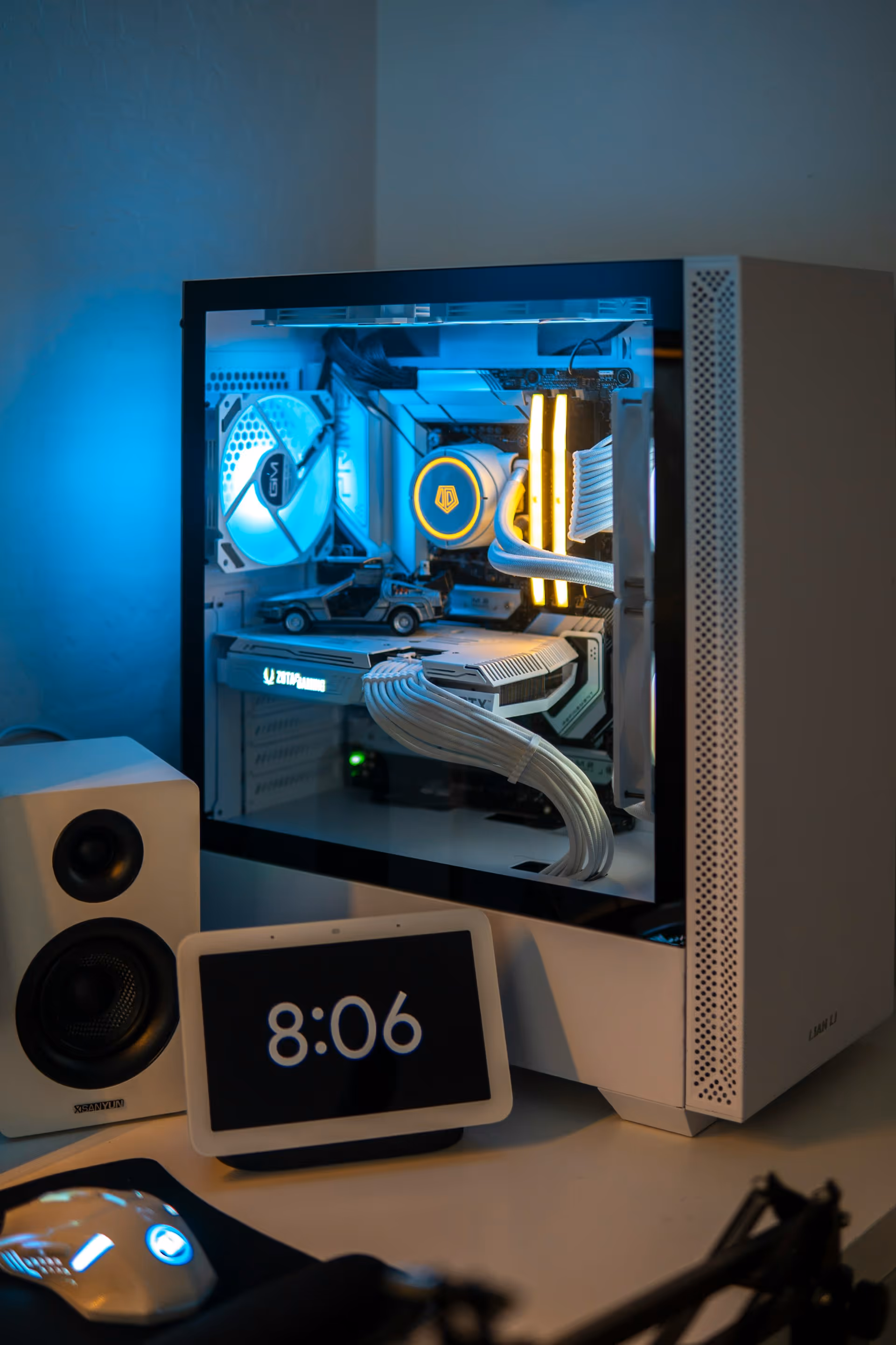 White gaming PC with illuminated interior components and miniature DeLorean model inside, next to a white speaker and a smart display showing 8:06.