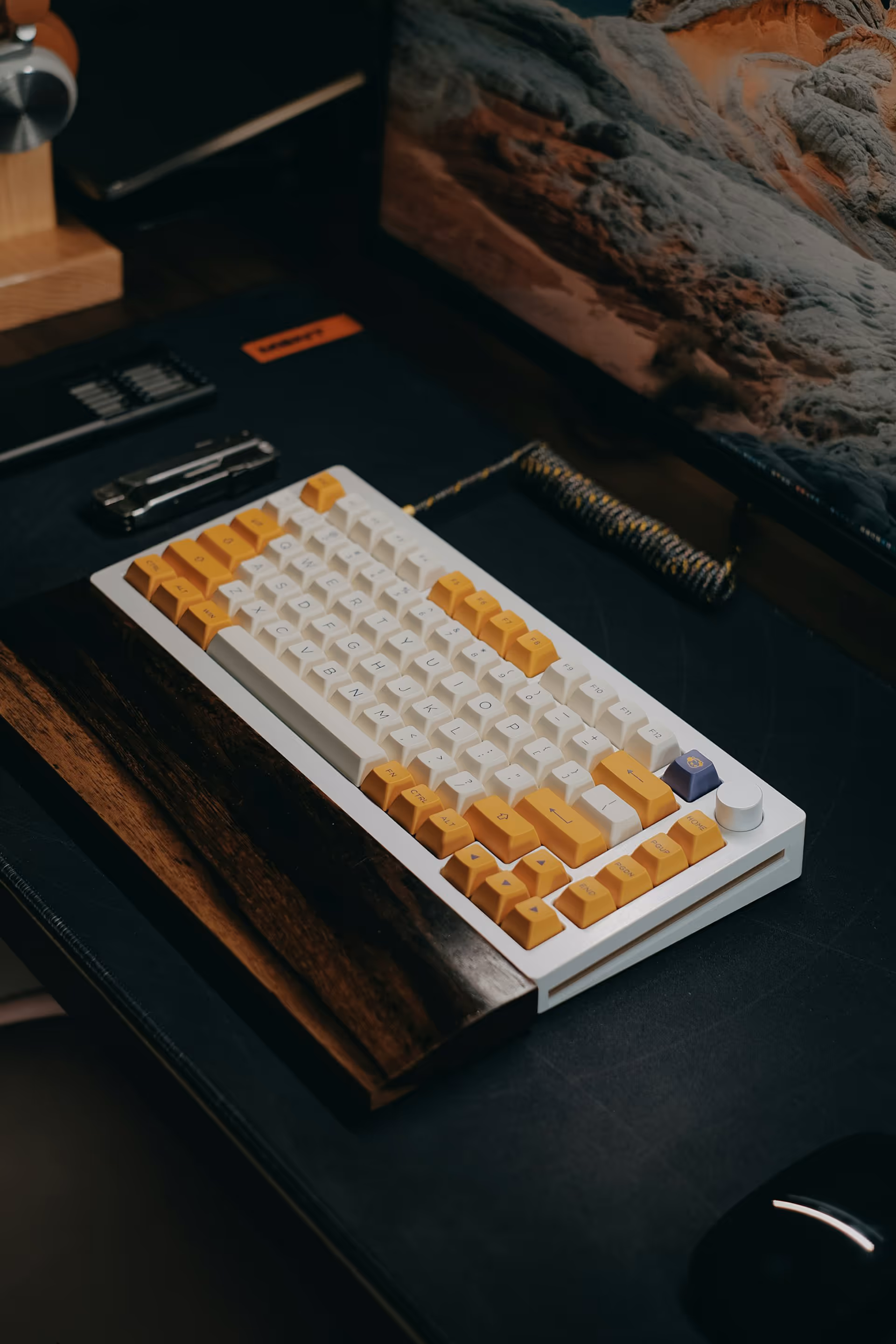 Mechanical keyboard with yellow and white keycaps and wooden wrist rest on a dark desk beside a monitor displaying a rocky landscape.