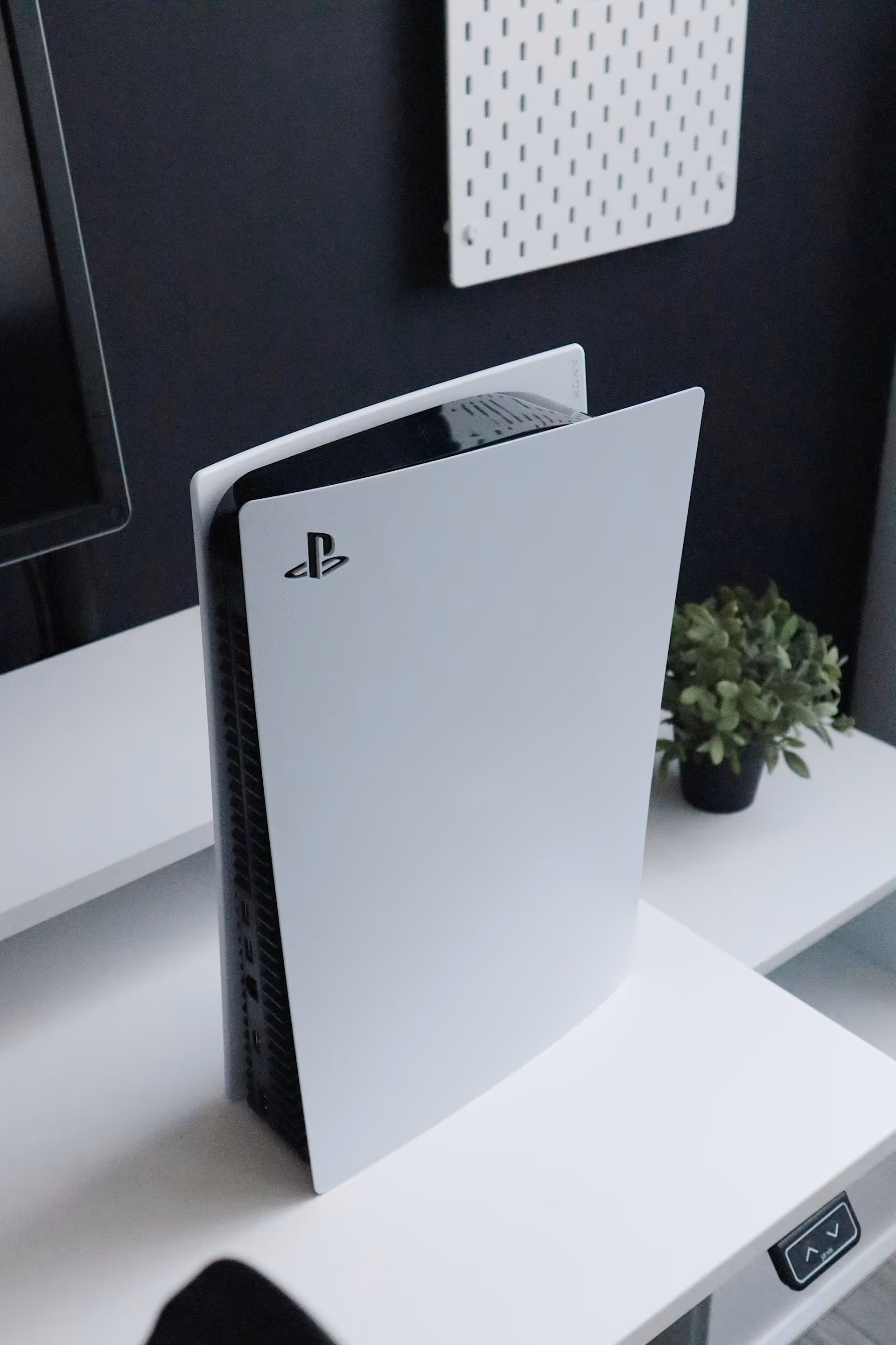 Sony PlayStation 5 console standing vertically on a white desk with a small potted plant in the background.