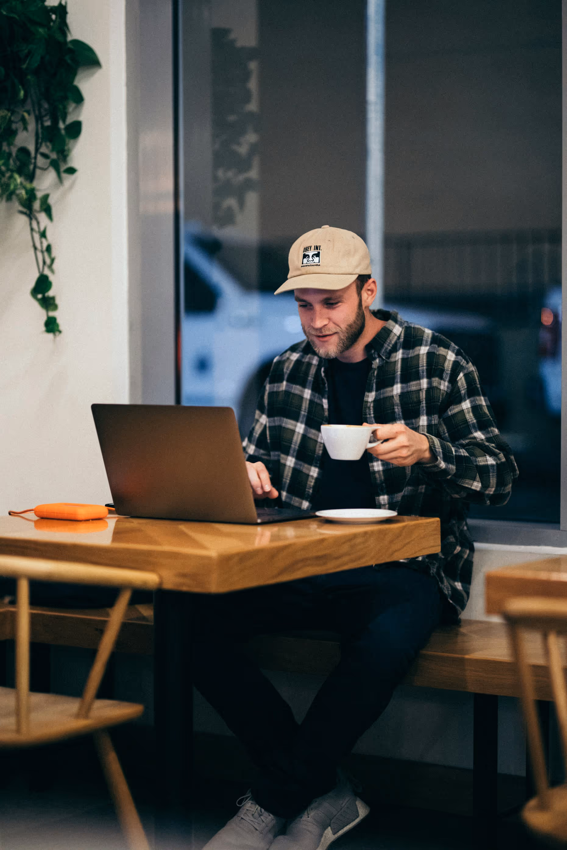 Man in a beige cap and plaid shirt sitting at a wooden table, using a laptop and holding a white coffee cup in a cozy café.