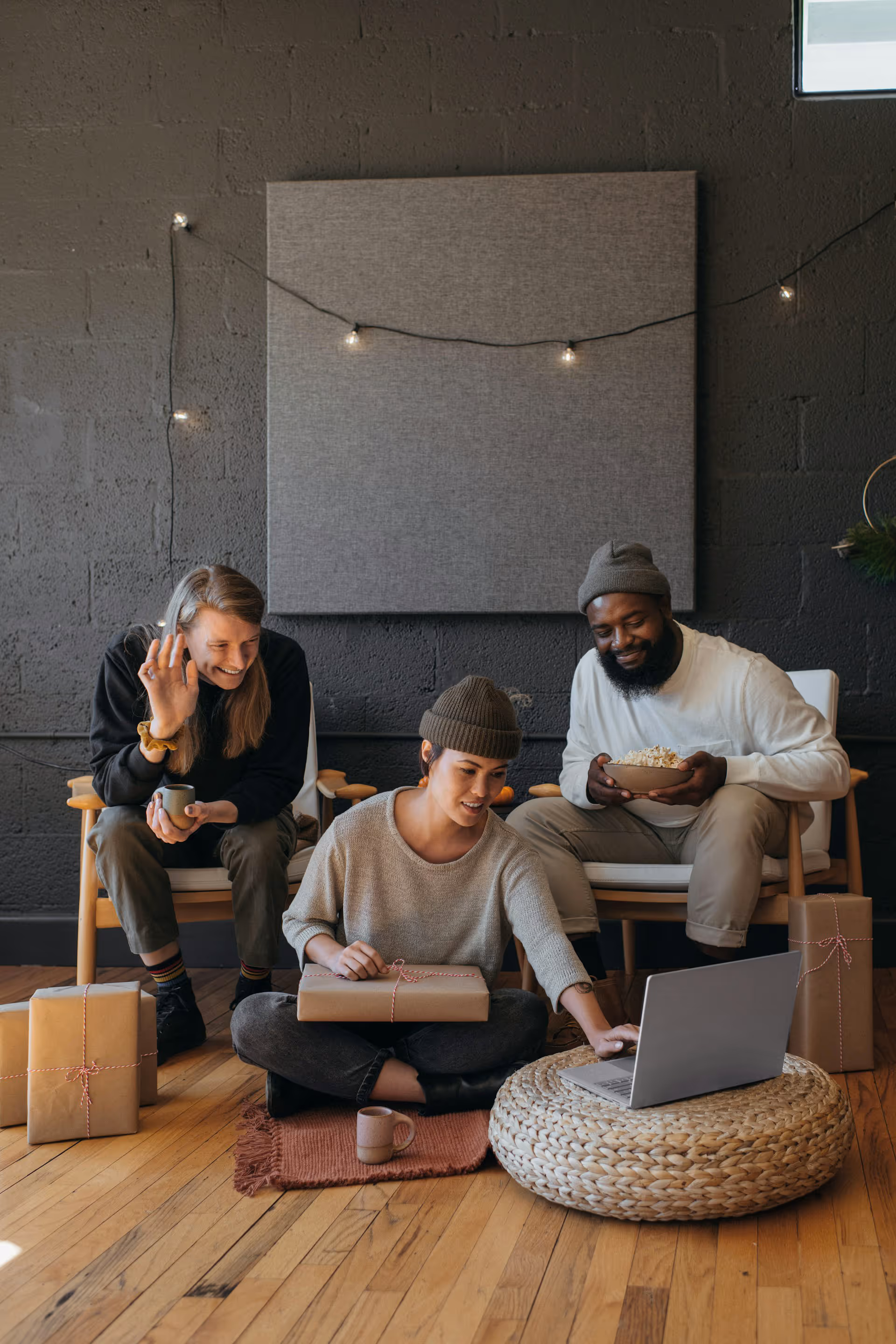 Three friends indoors with wrapped gifts, one waving, one using a laptop, and one holding popcorn.