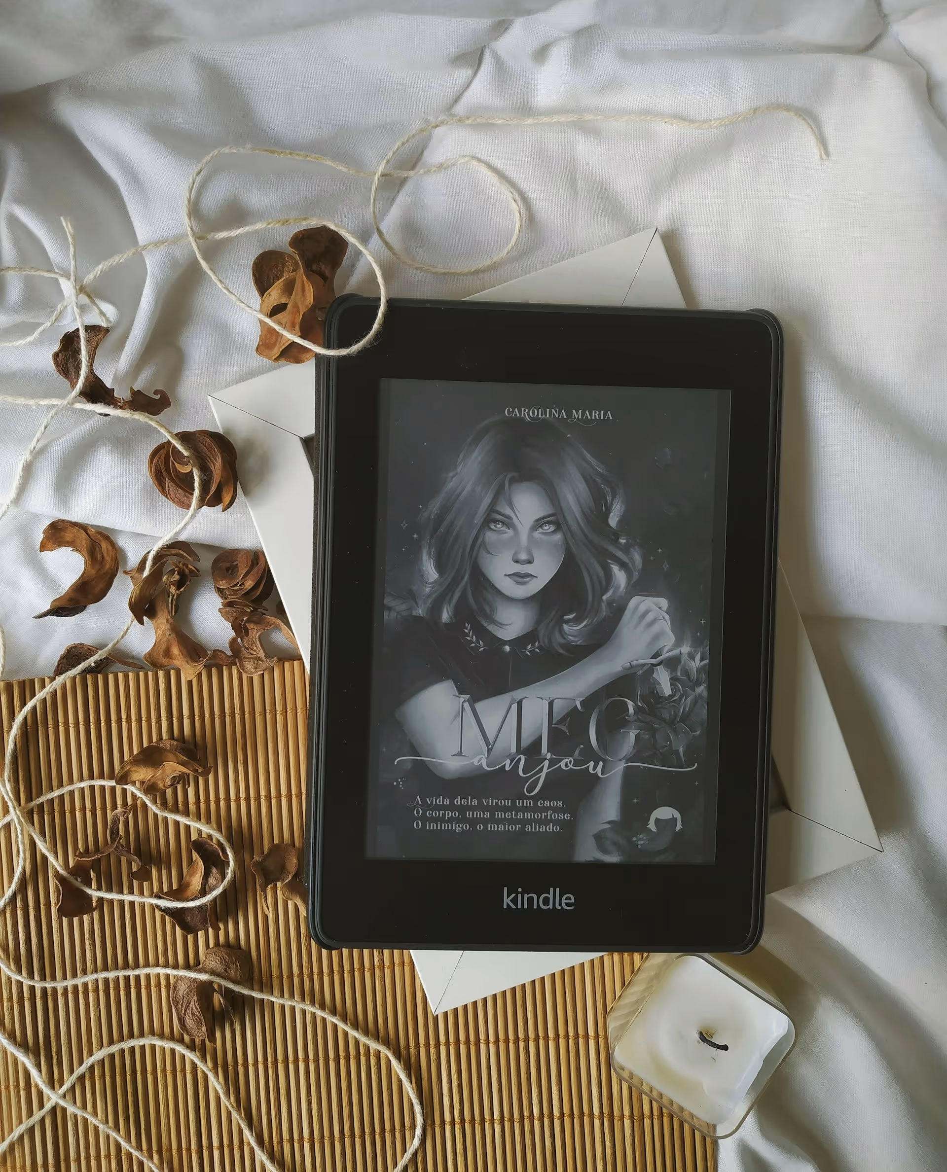 Kindle displaying a book cover with an illustrated woman, placed on white fabric with dried flower petals, string, and a white candle nearby.