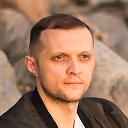 Portrait of a man with short brown hair and a trimmed beard wearing a black shirt, with a blurred natural rock background.