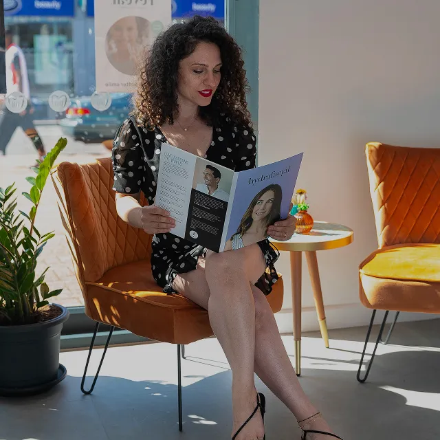 Woman with curly hair in a black polka dot dress sitting on an orange chair reading a magazine in a sunlit room.