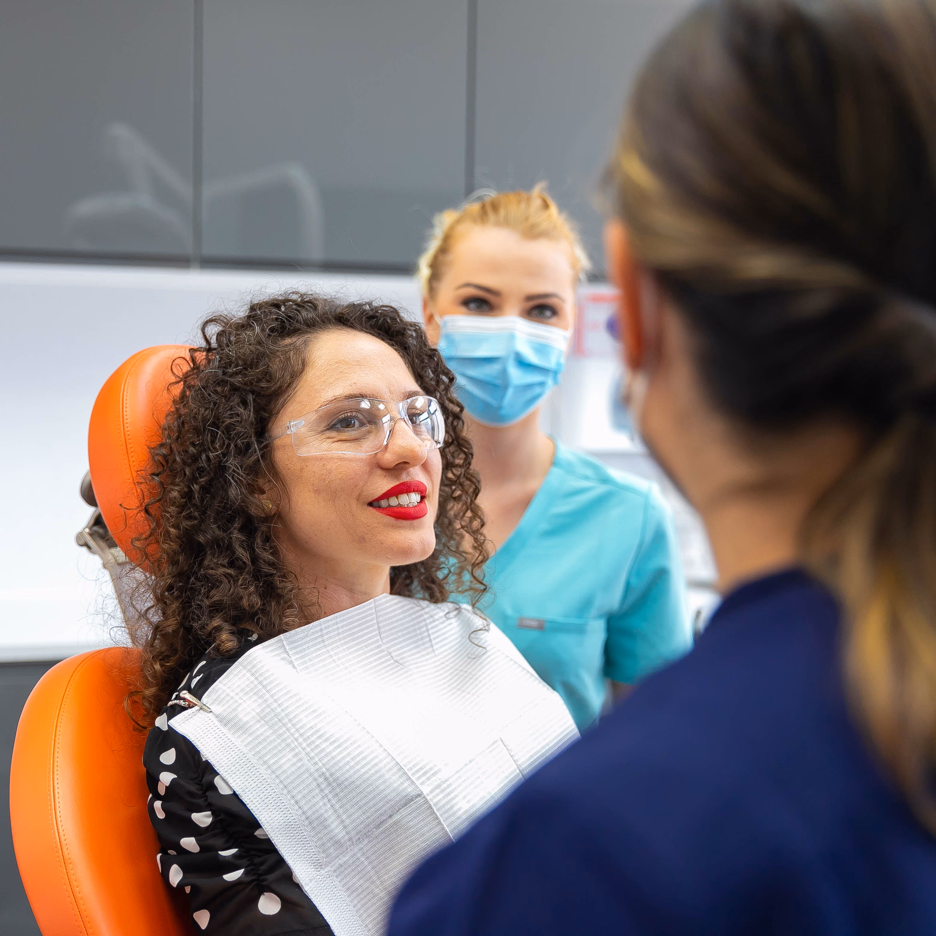 Woman patient wearing protective glasses sitting in an orange dental chair, smiling at a dentist with a masked dental assistant in the background.