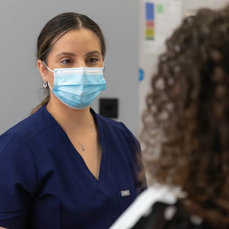 Healthcare worker in navy scrubs and a blue surgical mask talking to a patient with curly hair.