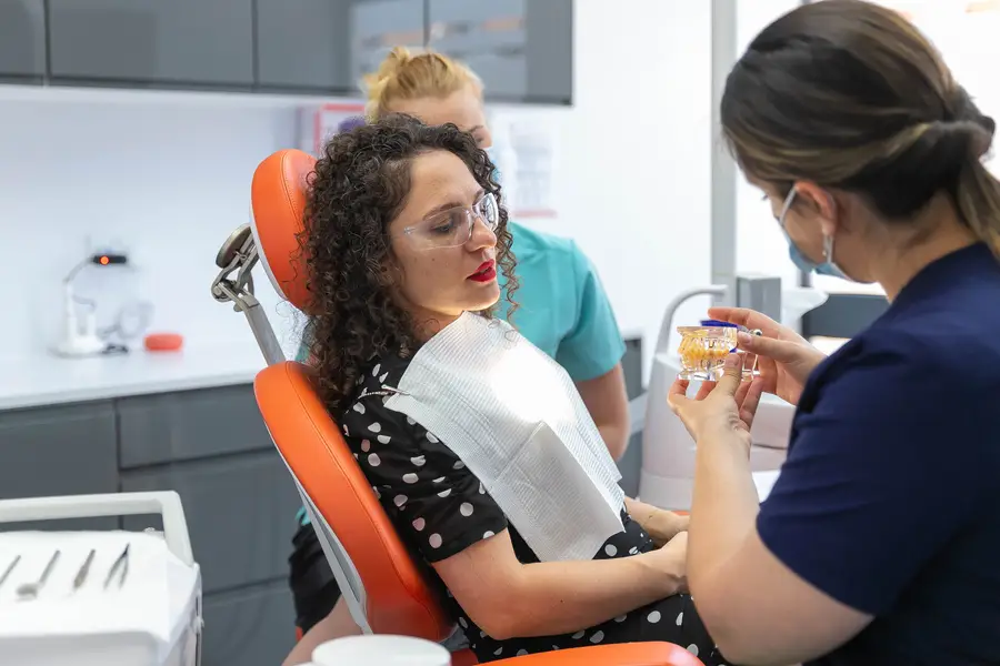 Dentist showing a dental model to a female patient seated in an orange dental chair in a clinic.