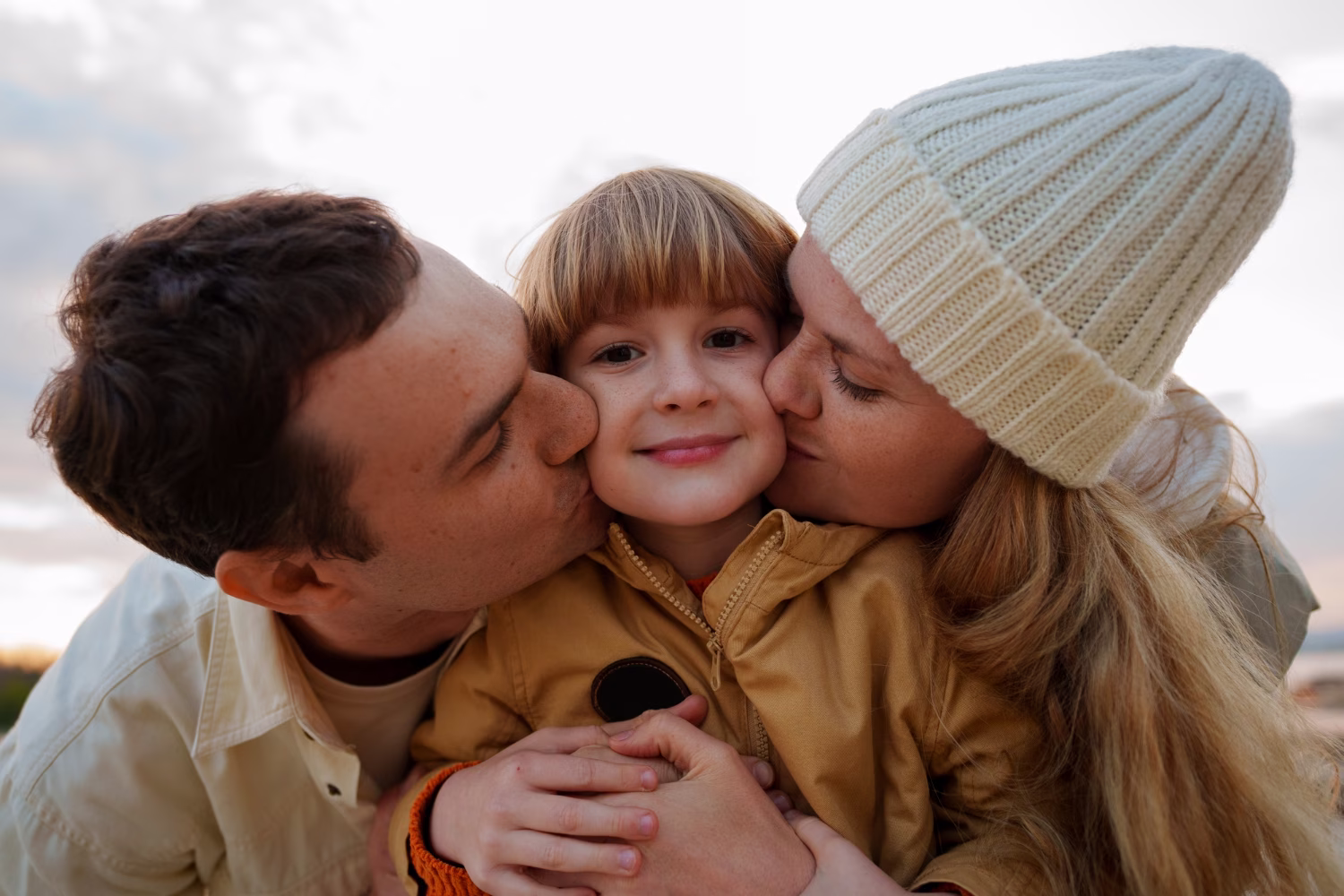 Child in a brown jacket smiling as two adults kiss their cheeks outdoors at sunset.