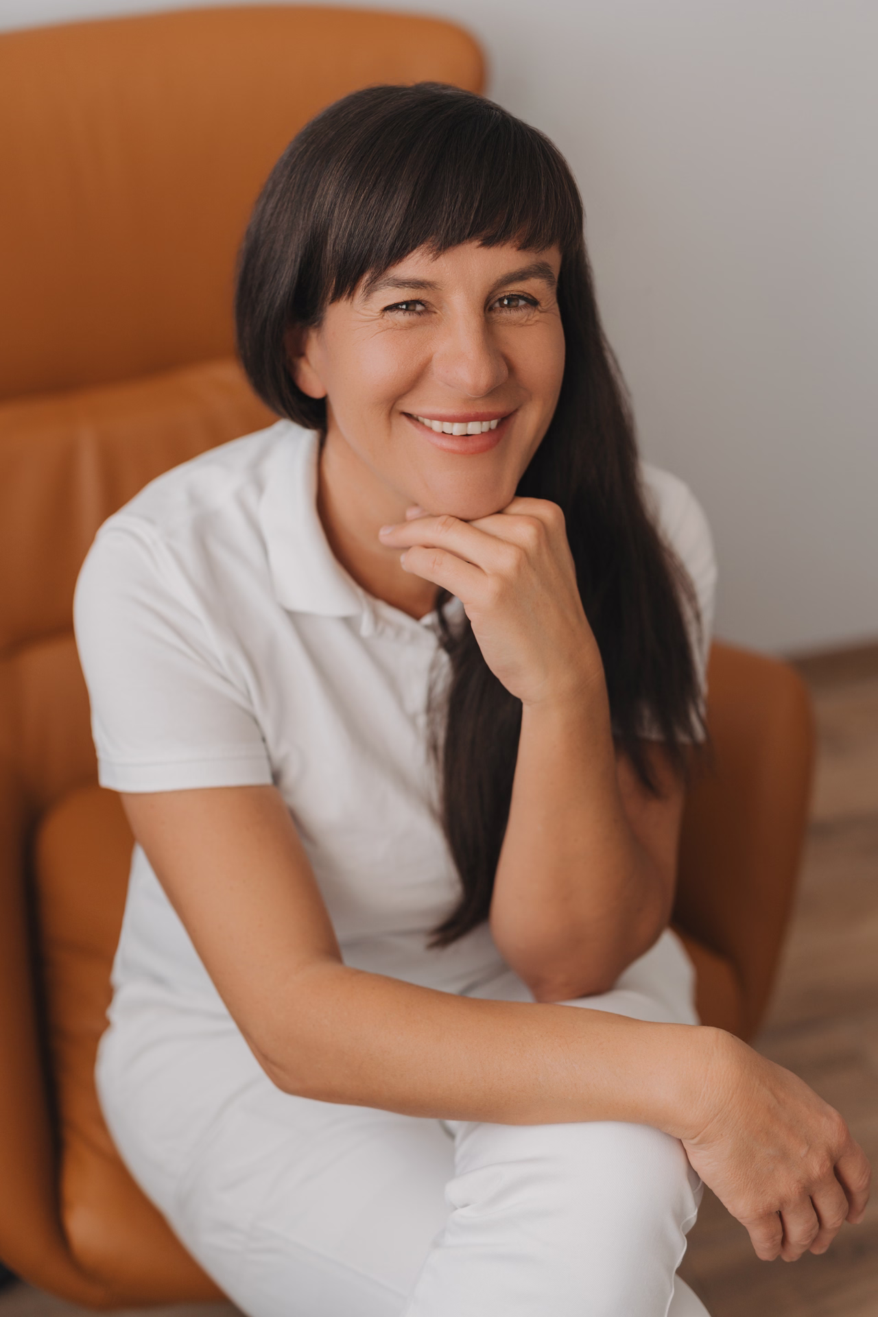 Smiling woman with dark hair wearing a white shirt sitting on a brown chair with hand on chin.