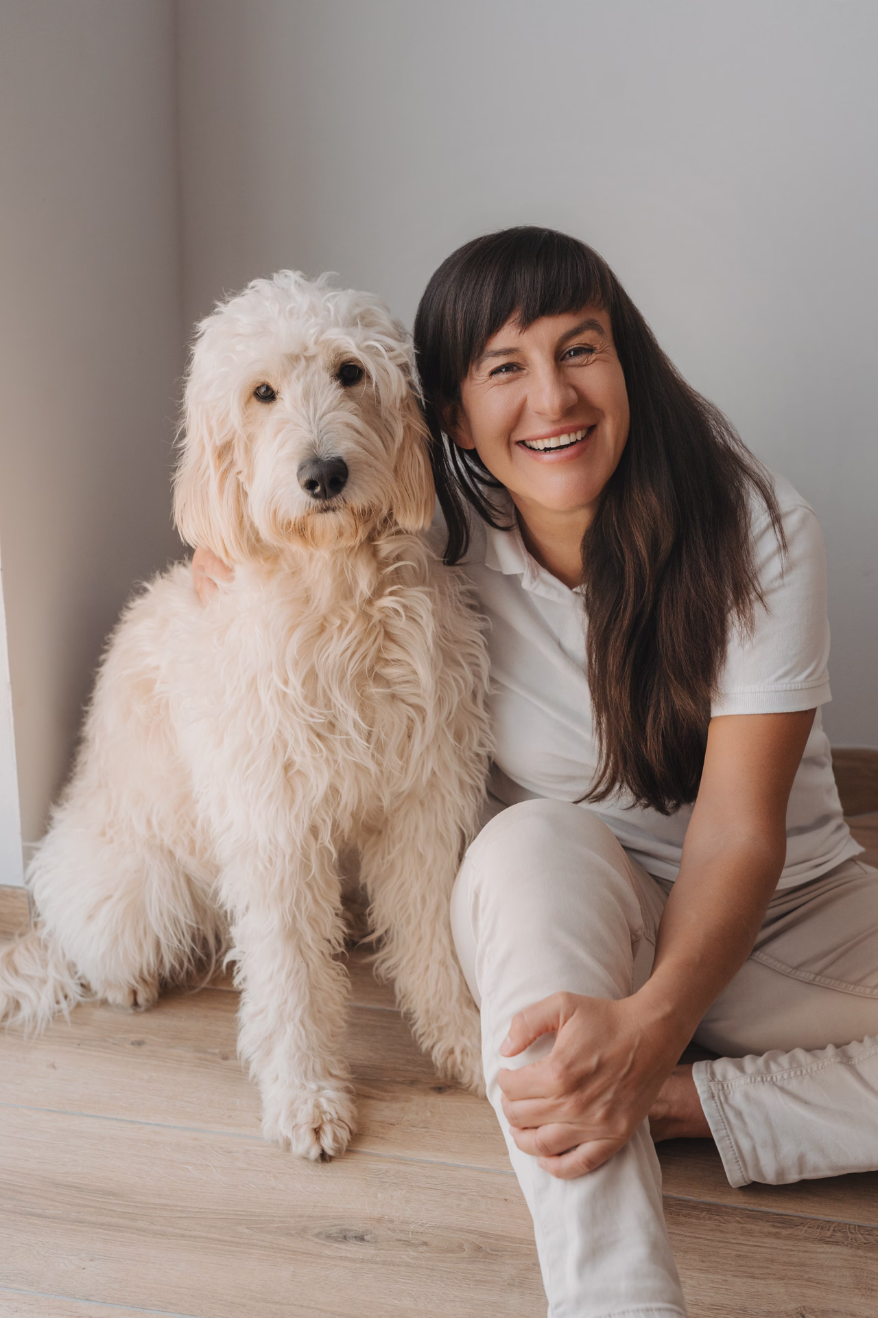 Smiling woman sitting on floor next to a large fluffy cream-colored dog indoors.