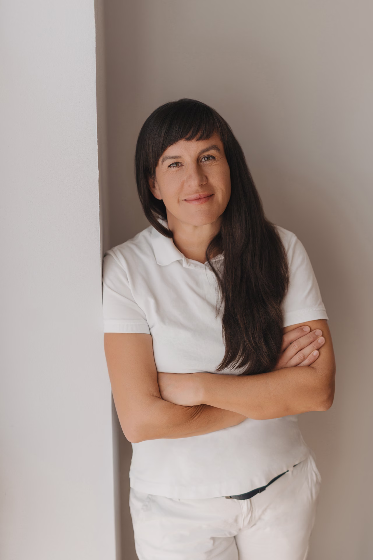 Smiling woman with long dark hair wearing white polo and pants, standing with arms crossed.