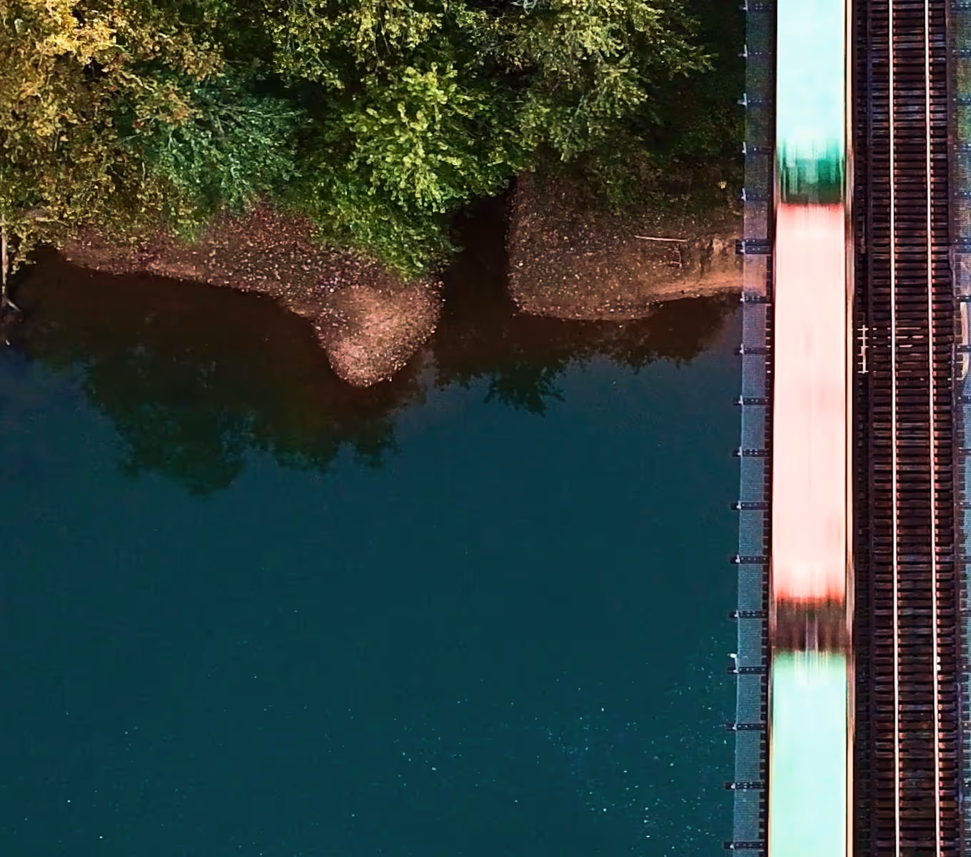 Aerial view of a blurred train moving on railway tracks beside a riverbank with green trees.