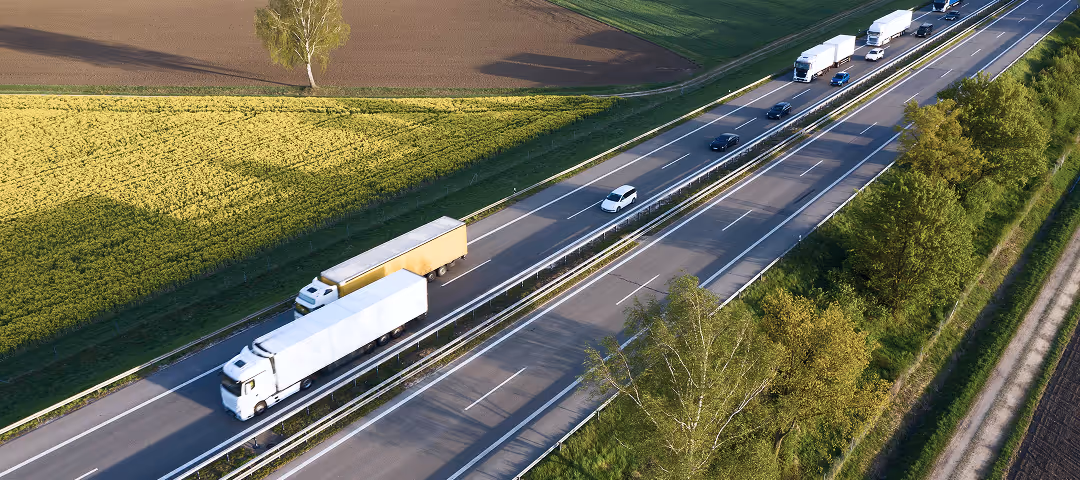 Aerial view of a highway with trucks and cars driving between fields and trees on a sunny day.