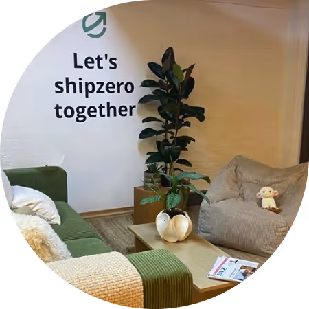 Cozy lounge area with a green sofa, a beige bean bag chair, a coffee table with magazines, and a tall plant against a wall with text 'Let's shipzero together'.