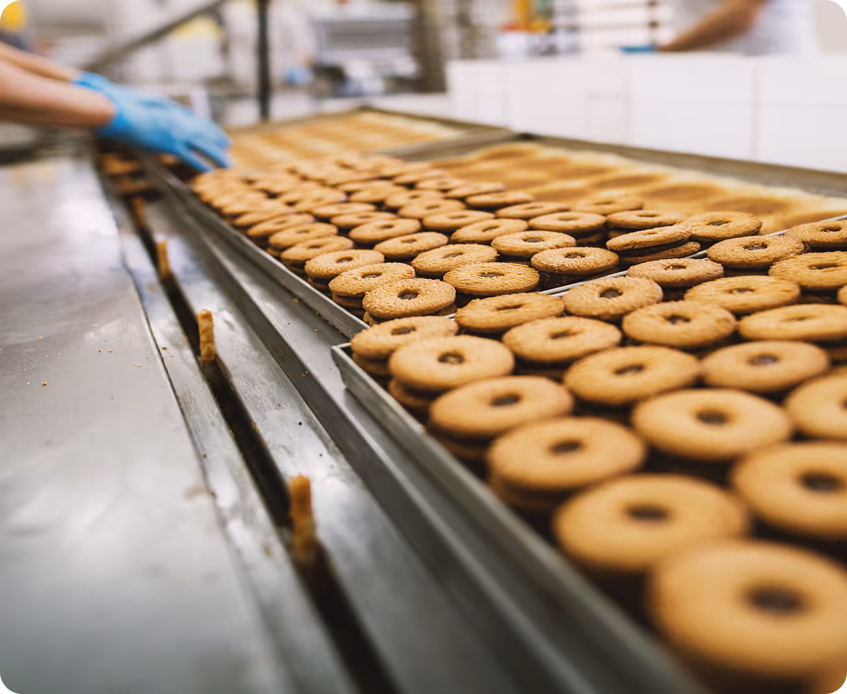 Rows of round cookies with holes in the center on trays in a bakery production line with a worker wearing blue gloves in the background.