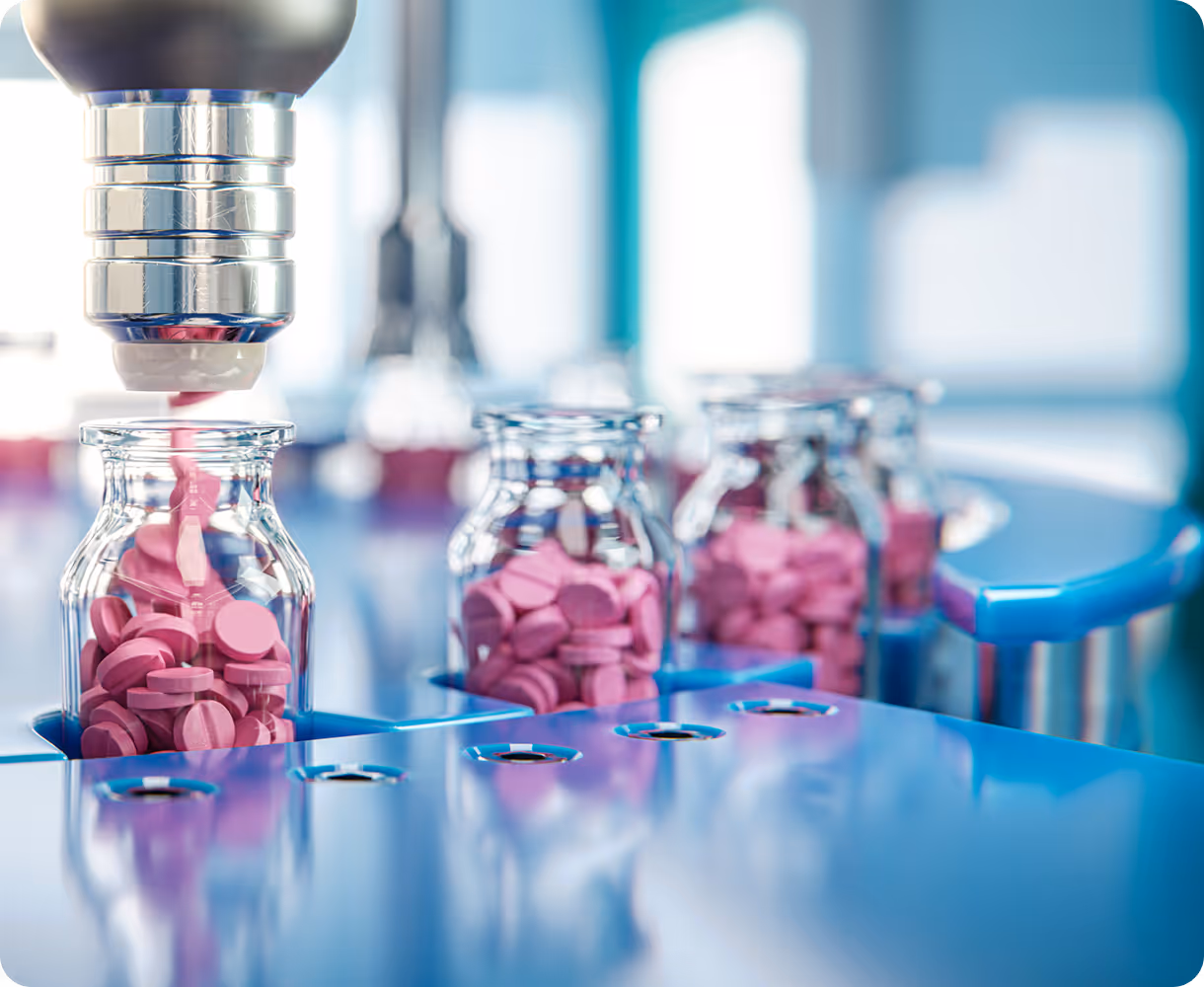 Robotic arm placing pink round tablets into glass vials on a pharmaceutical production line.