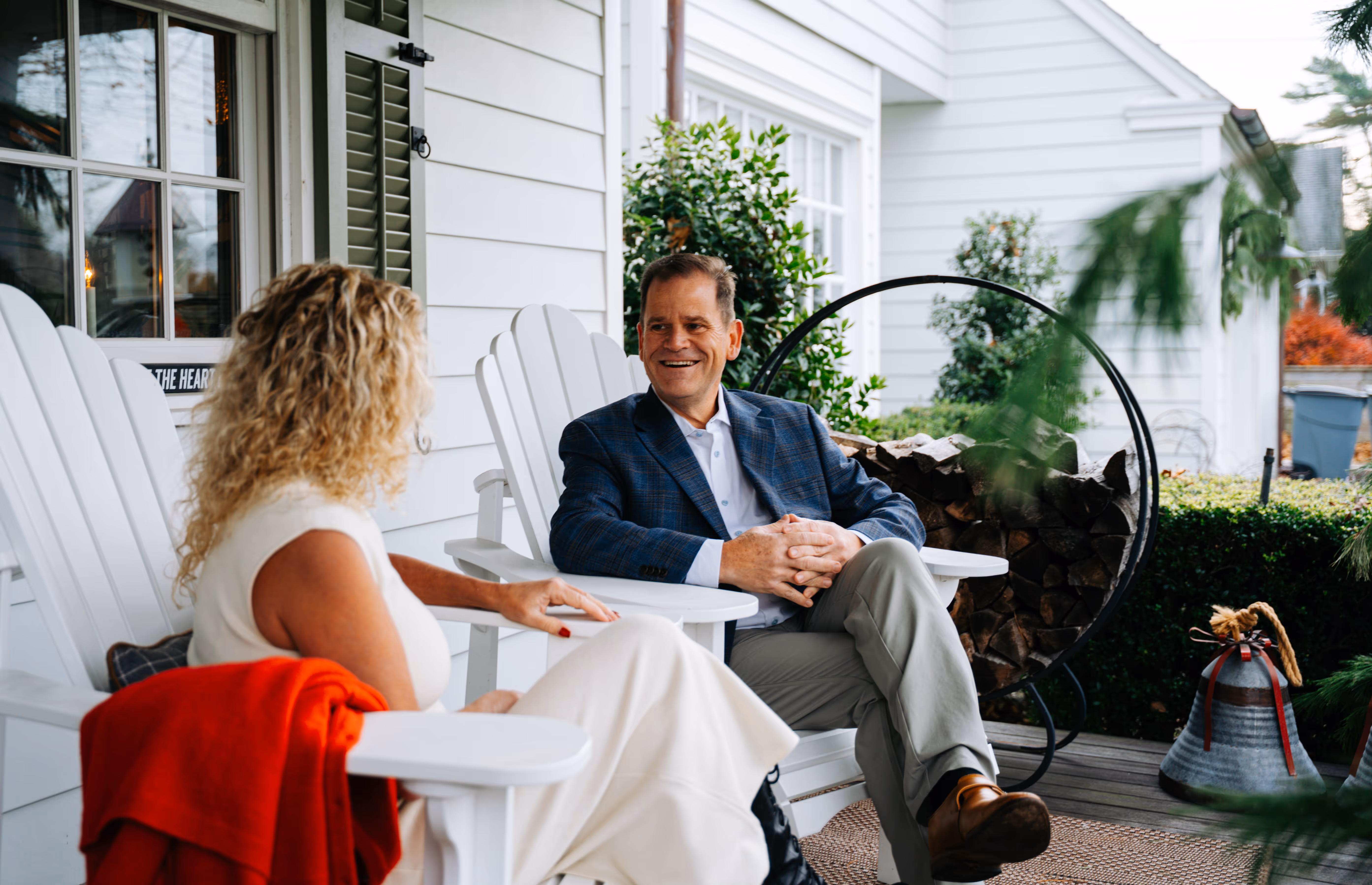Man in a blue blazer and khaki pants and woman in a white dress sitting and talking on a porch with white Adirondack chairs and stacked firewood in a ring holder.