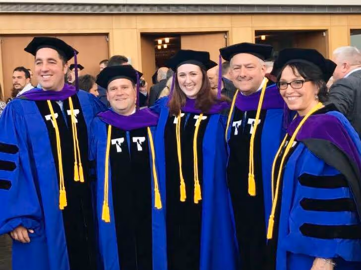 Five graduates wearing blue academic gowns, purple hoods, and black caps, smiling and standing indoors at graduation.