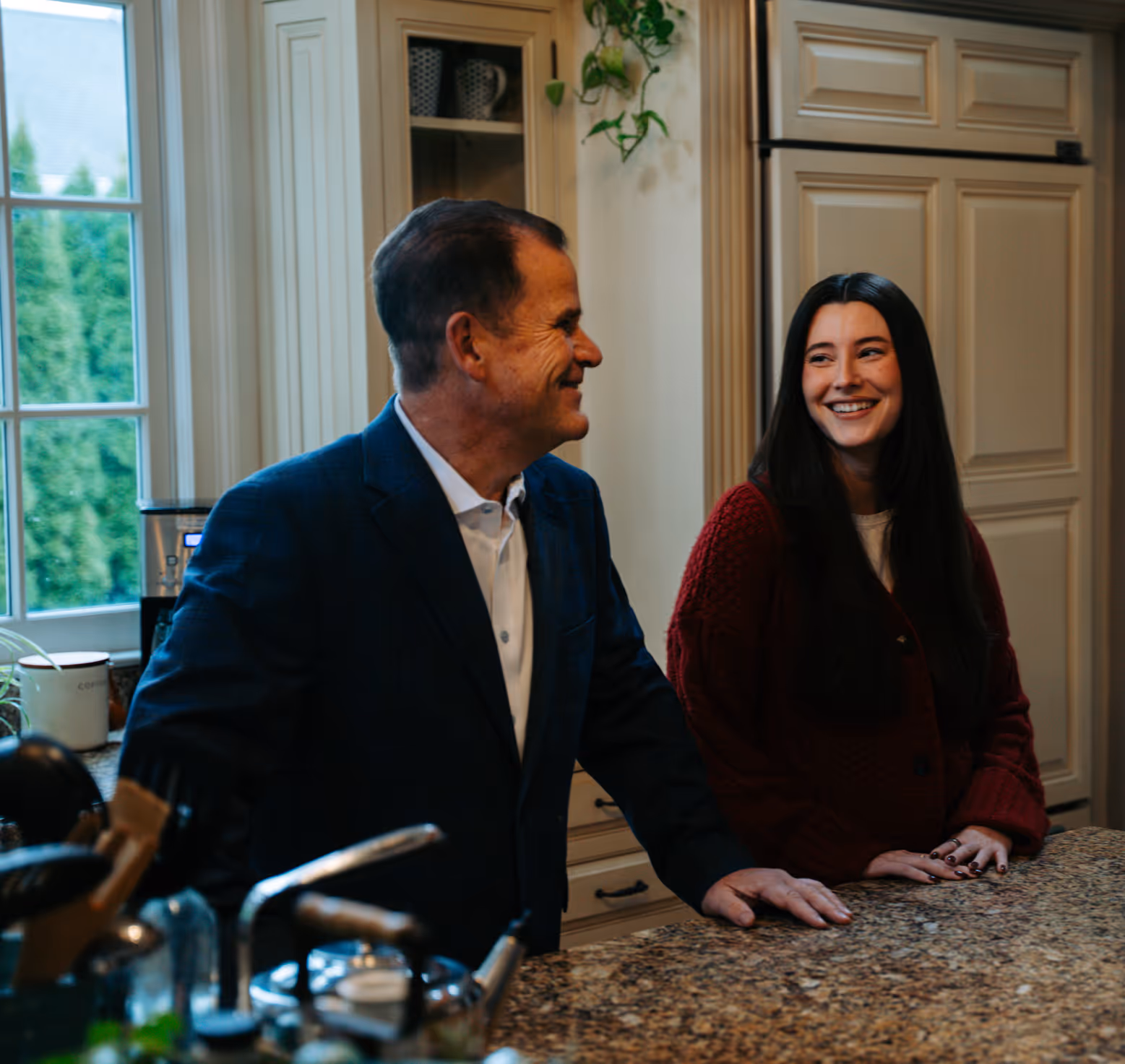Man in a dark suit and white shirt smiling at a woman with long dark hair in a red sweater in a kitchen.