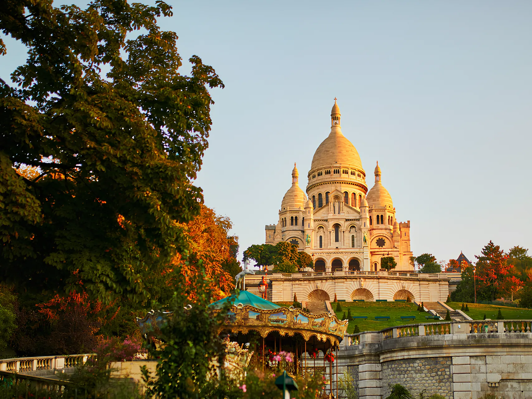 La basilique du Sacré-Cœur illuminée par le soleil couchant avec un manège coloré et des arbres verts en premier plan.
