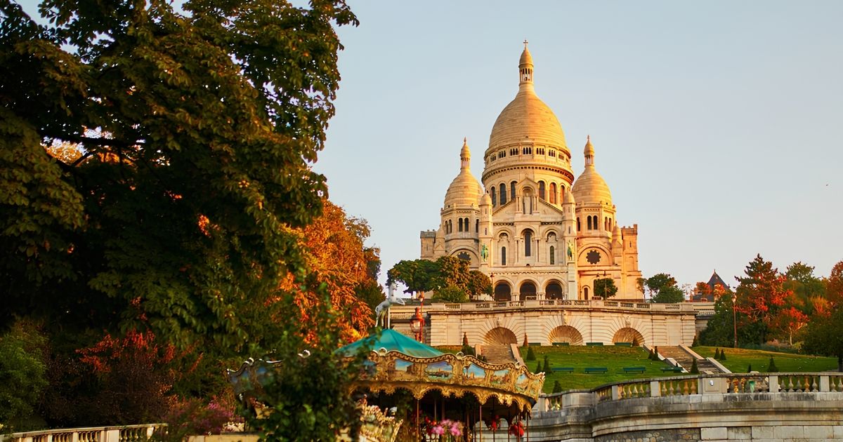 La basilique du Sacré-Cœur éclairée par le soleil couchant, avec un carrousel coloré et des arbres aux feuilles d'automne au premier plan.