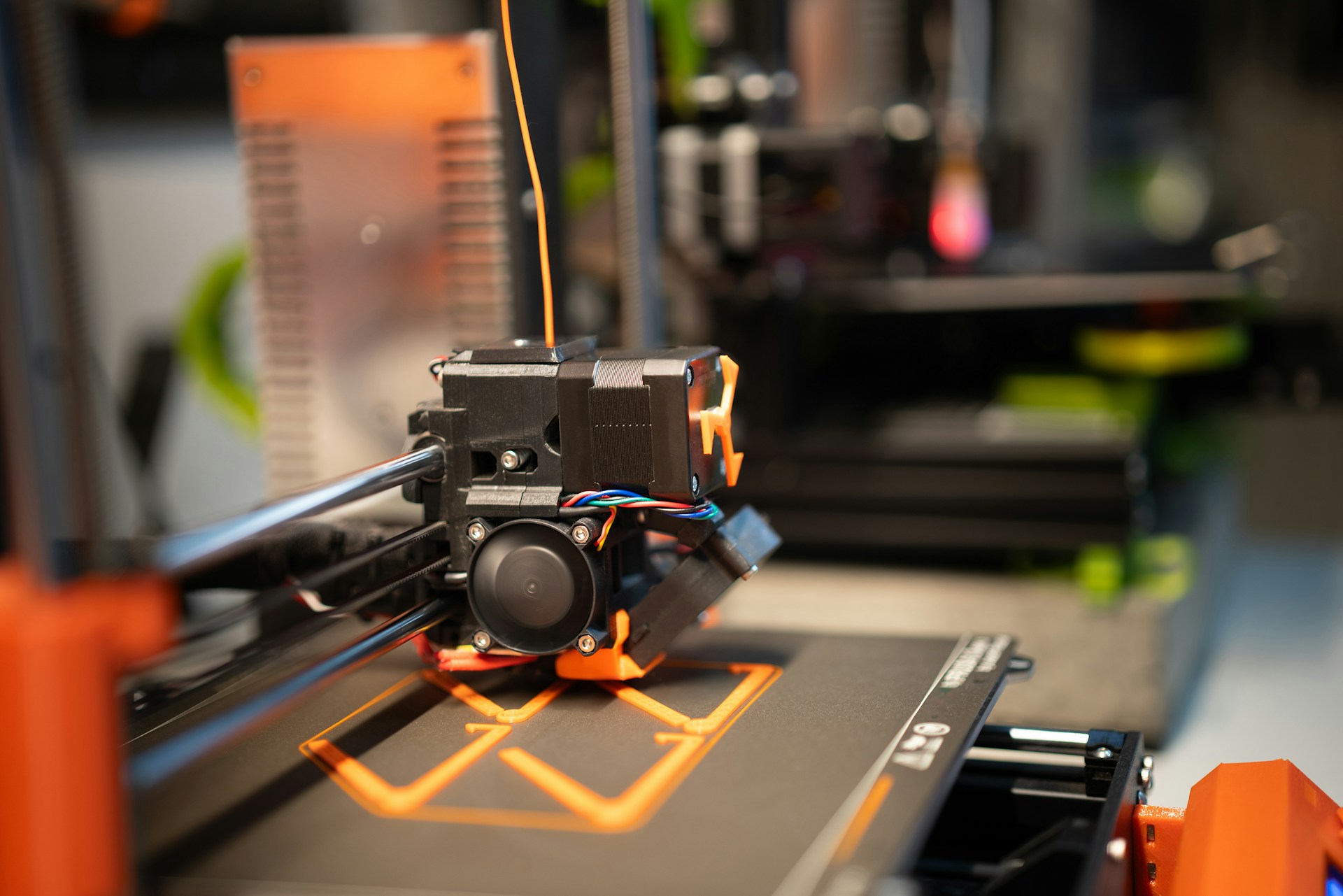 Close-up of a 3D printer nozzle extruding orange filament to print a shape on the printer bed.