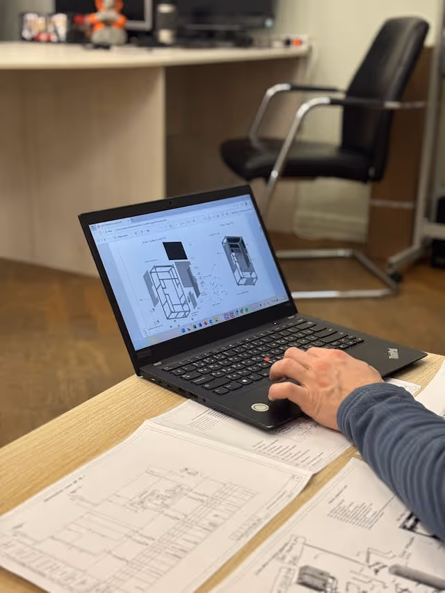 Person working on a laptop displaying architectural 3D plans, with printed technical drawings on the table.