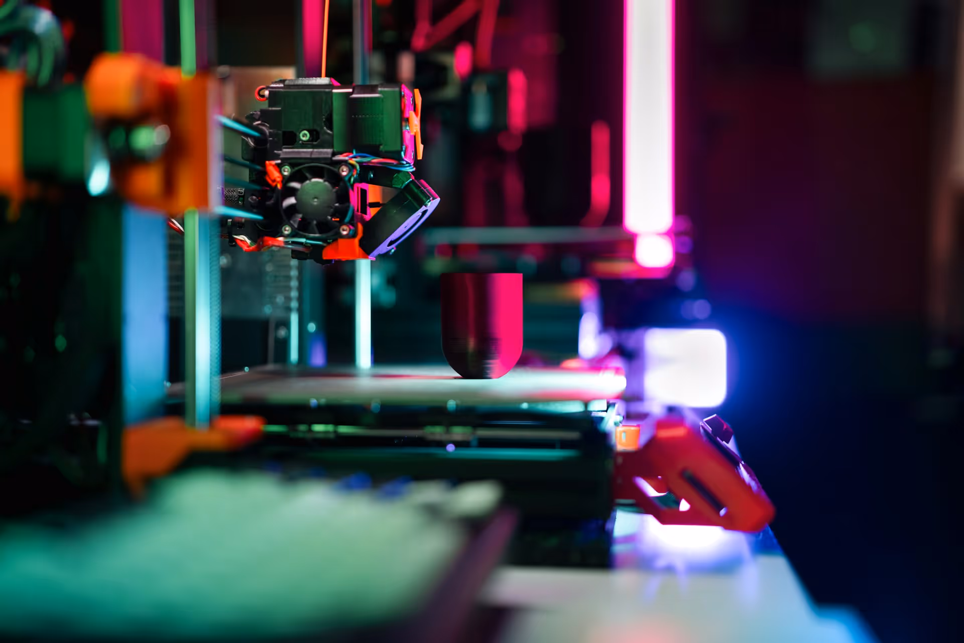 Close-up of a 3D printer nozzle printing a cylindrical object with colorful LED lights in the background.