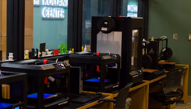 A row of 3D printers on wooden desks inside a modern workshop with chairs and a window showing the Rolland Center sign.