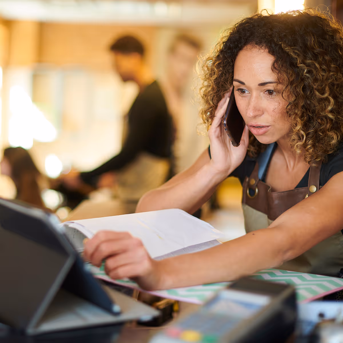 Woman in apron talking on phone while working at a counter in a busy cafe.