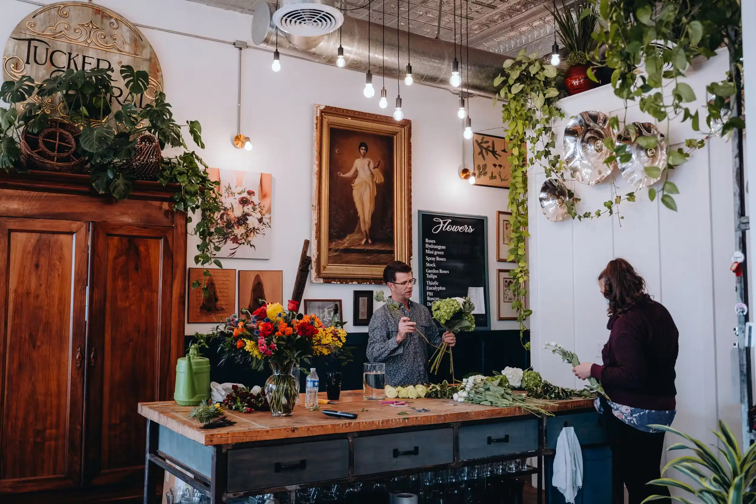 A florist arranges flowers at a worktable in Tucker Floral, a plant-filled shop with vintage art, hanging greenery, and a chalkboard flower menu.
