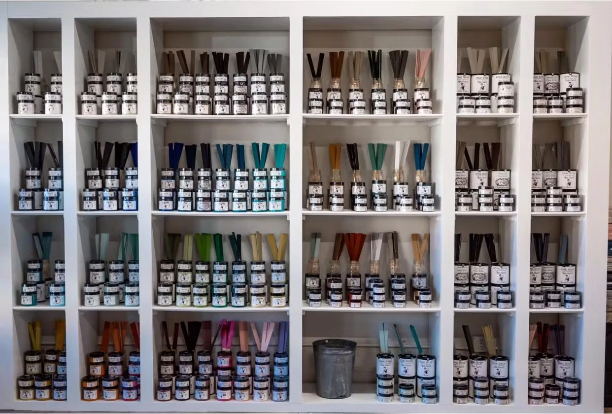 A white shelving unit displaying rows of paint jars with color-coded sample sticks in a range of hues.