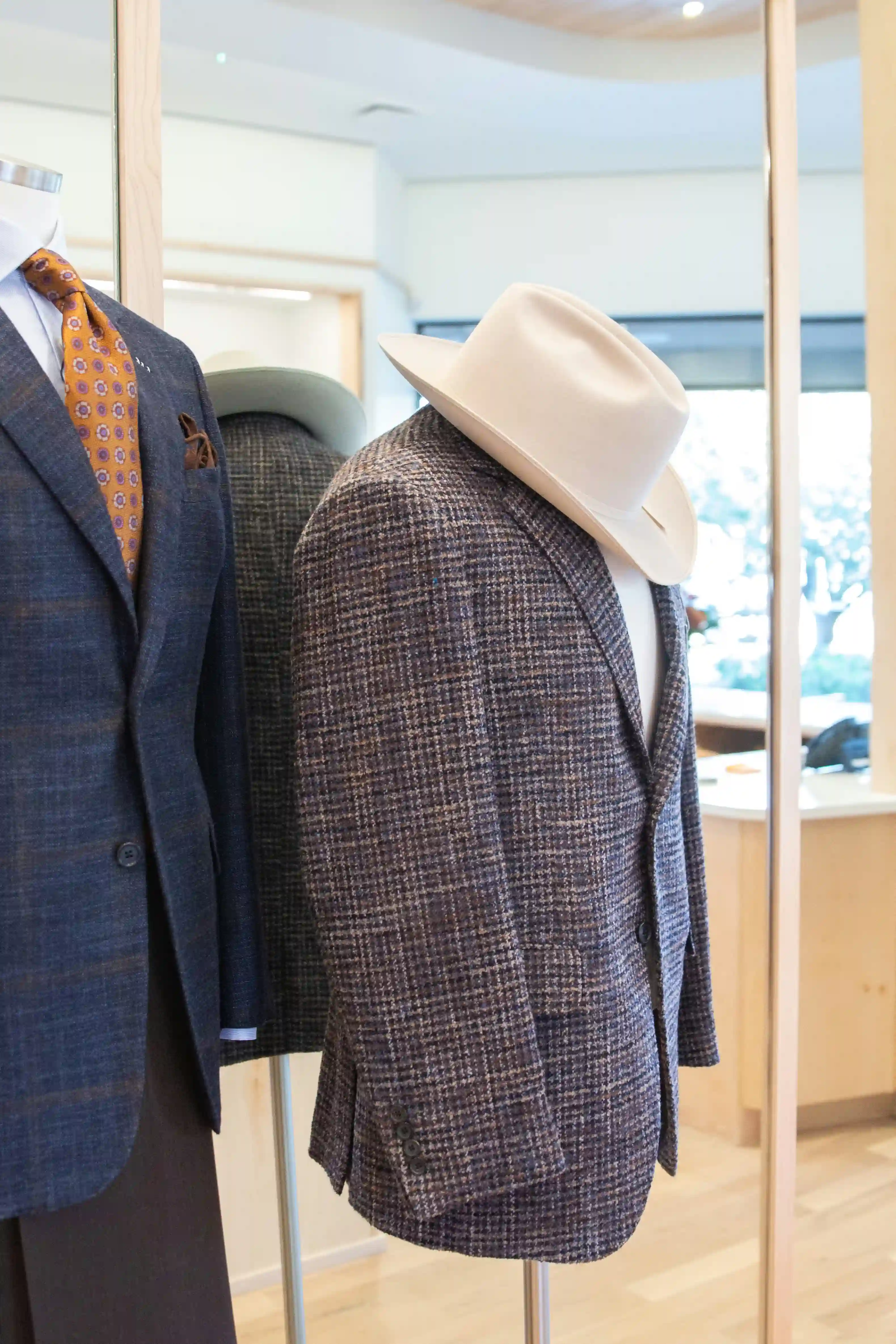 Mannequins displaying tailored sport coats in a menswear shop, one topped with a cream cowboy hat and the other paired with an orange patterned tie.