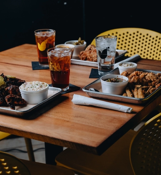 A wooden table set with trays of fried chicken, mashed potatoes, and sides, alongside glasses of iced tea and water.
