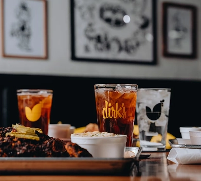 Close-up of a glass of iced tea branded "dirk's" on a table with food and other drinks, framed art blurred in the background.