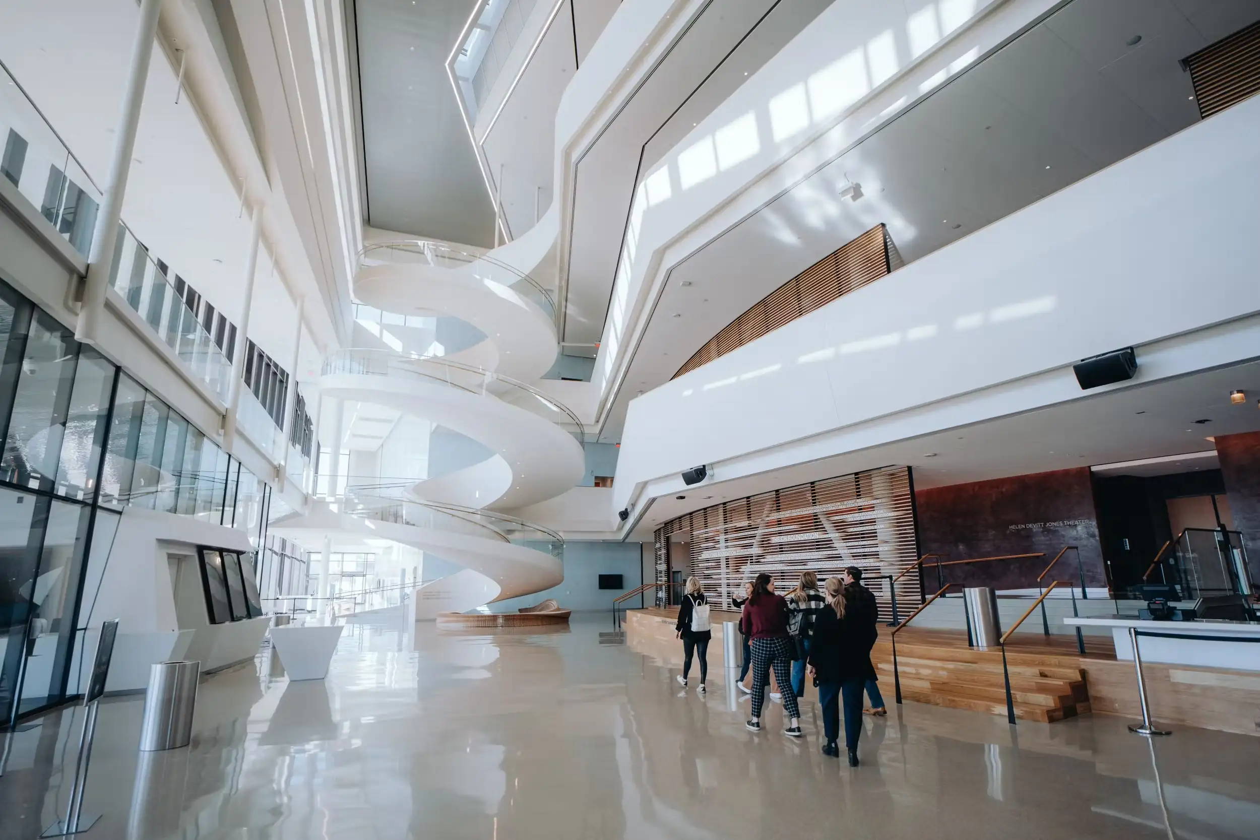 A group of people walk through a bright, modern lobby with a sculptural white spiral staircase and floor-to-ceiling windows.