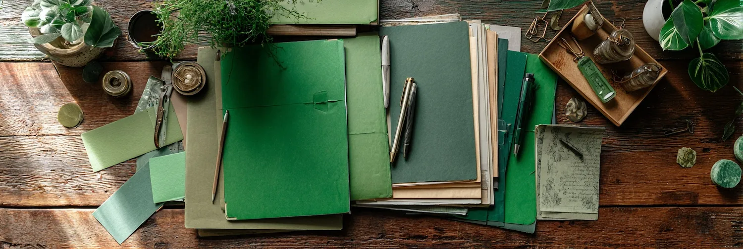 Top view of a rustic wooden table with green notebooks, pens, pencils, plants, and glass jars arranged neatly.