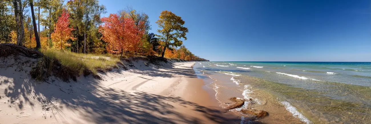 Sandy shoreline with clear waves and trees with autumn foliage under a blue sky.