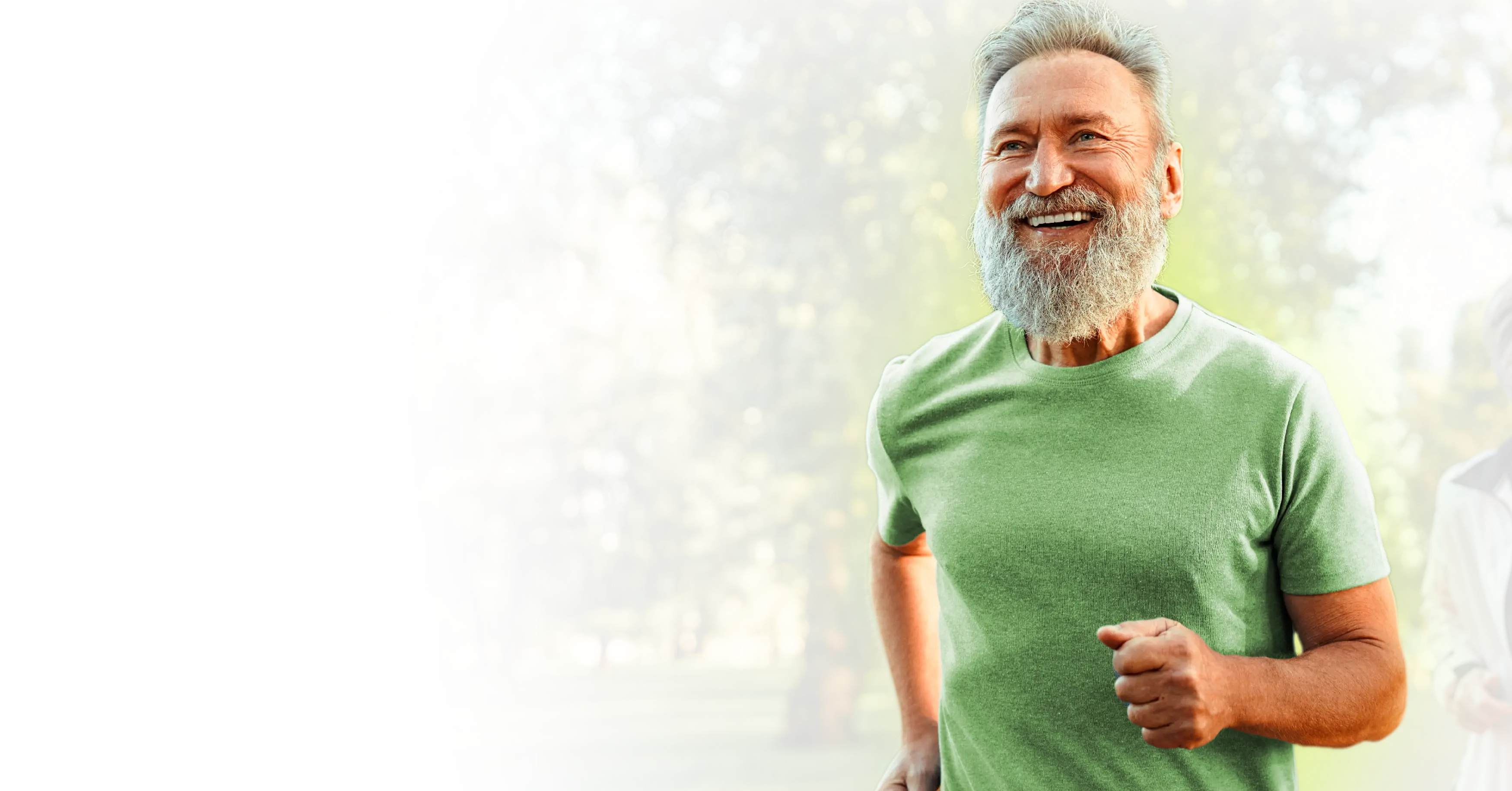 Smiling elderly man with gray beard jogging outdoors in a green t-shirt with trees in the background.