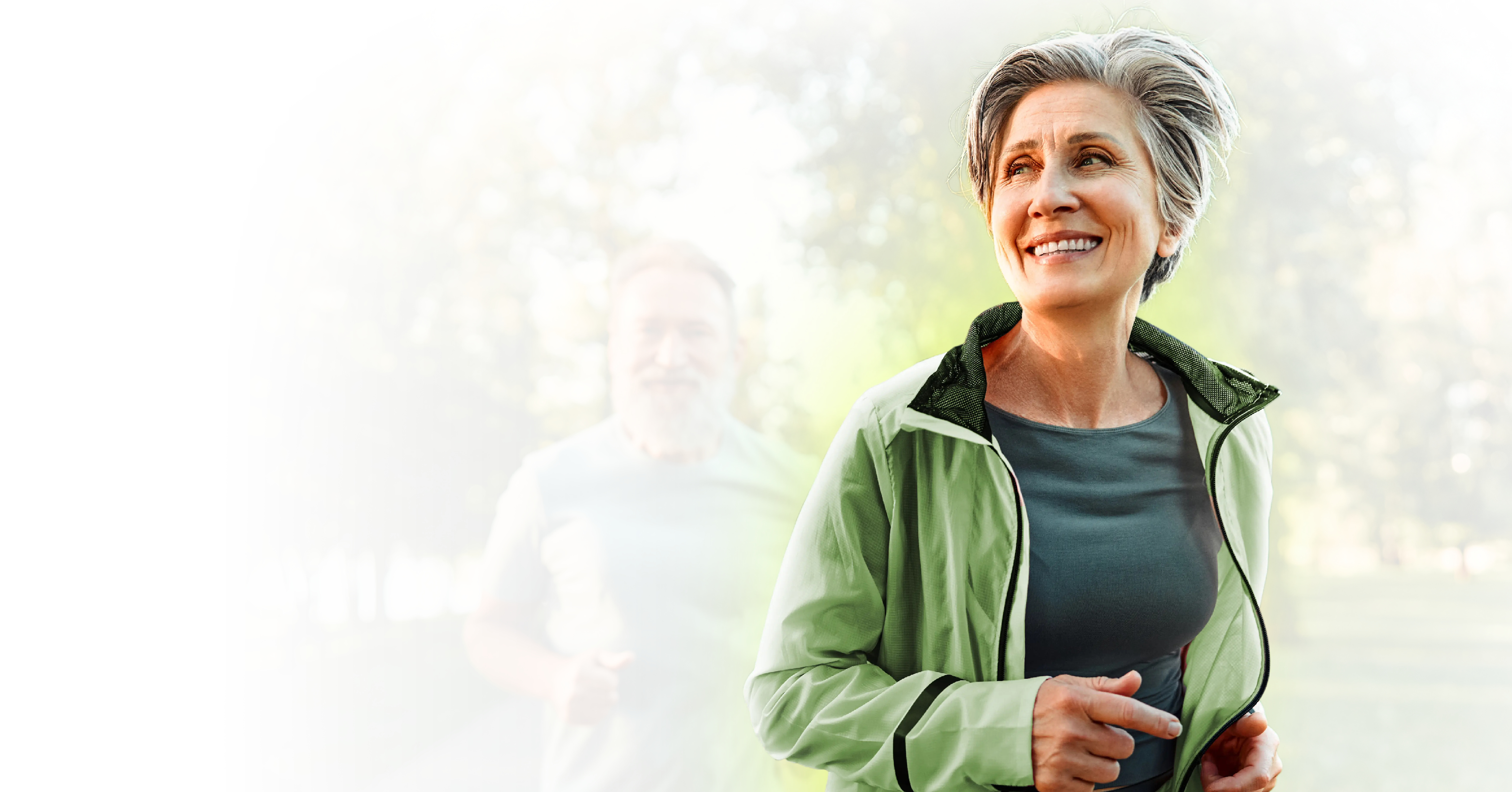 Smiling senior woman jogging outdoors in a green jacket with a bearded man running behind her on a tree-lined path.