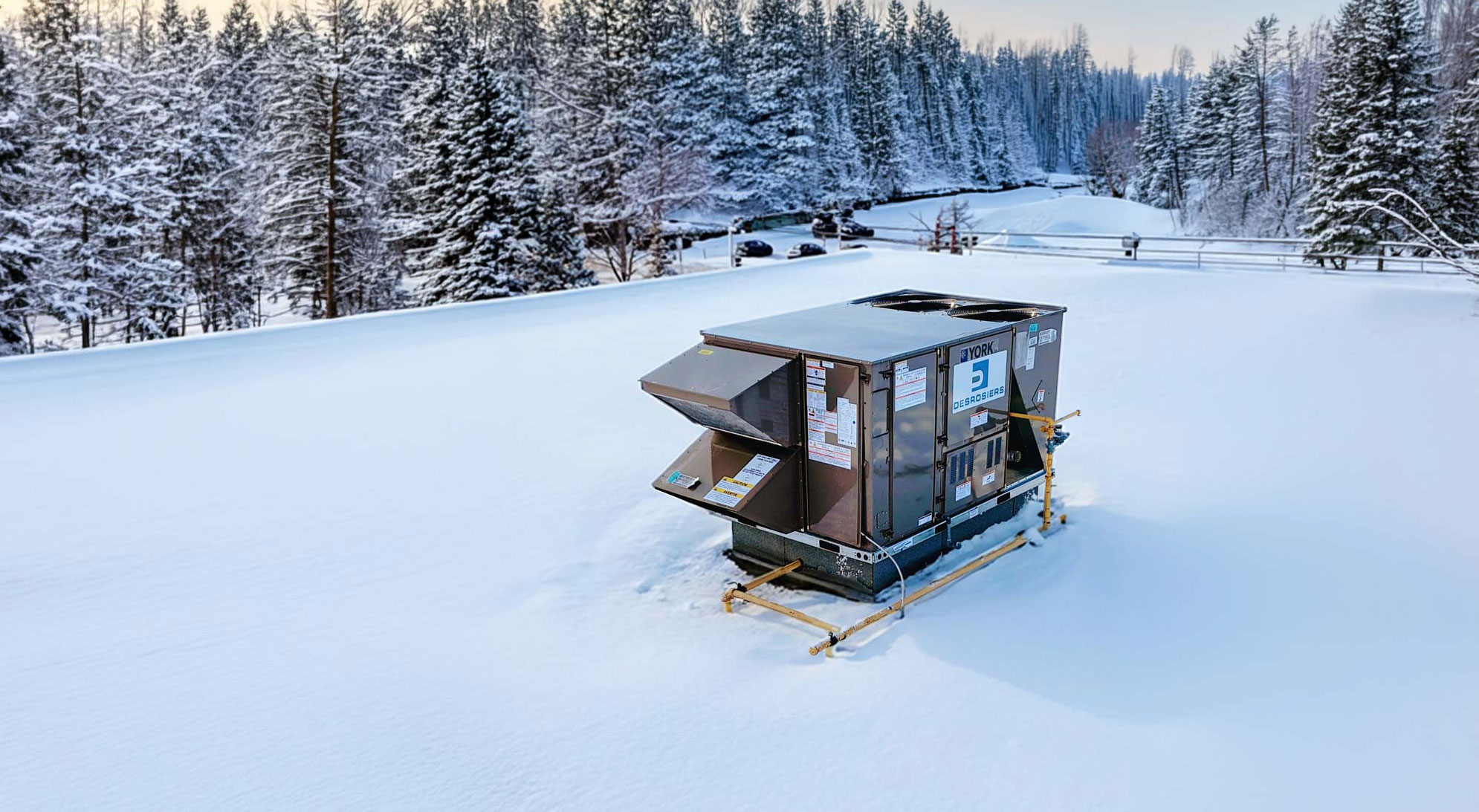 Unité de climatisation YORK installée sur un toit plat couvert de neige, avec une épaisse couche de neige sur toute la surface et des arbres enneigés en arrière-plan.