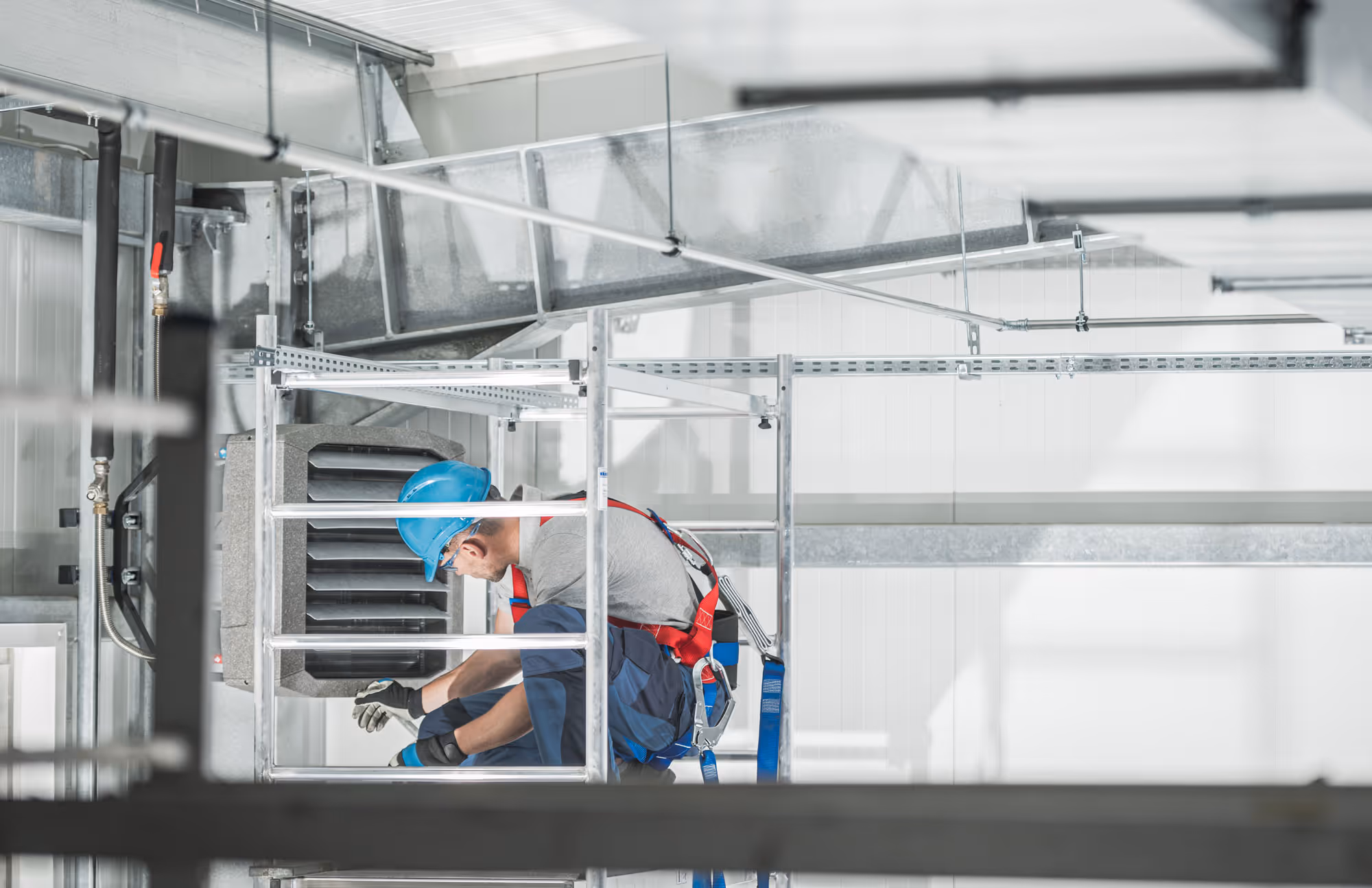Travailleur portant un casque bleu et un harnais de sécurité effectuant l'entretien ou l'installation d'un système de ventilation industriel dans un espace intérieur moderne.