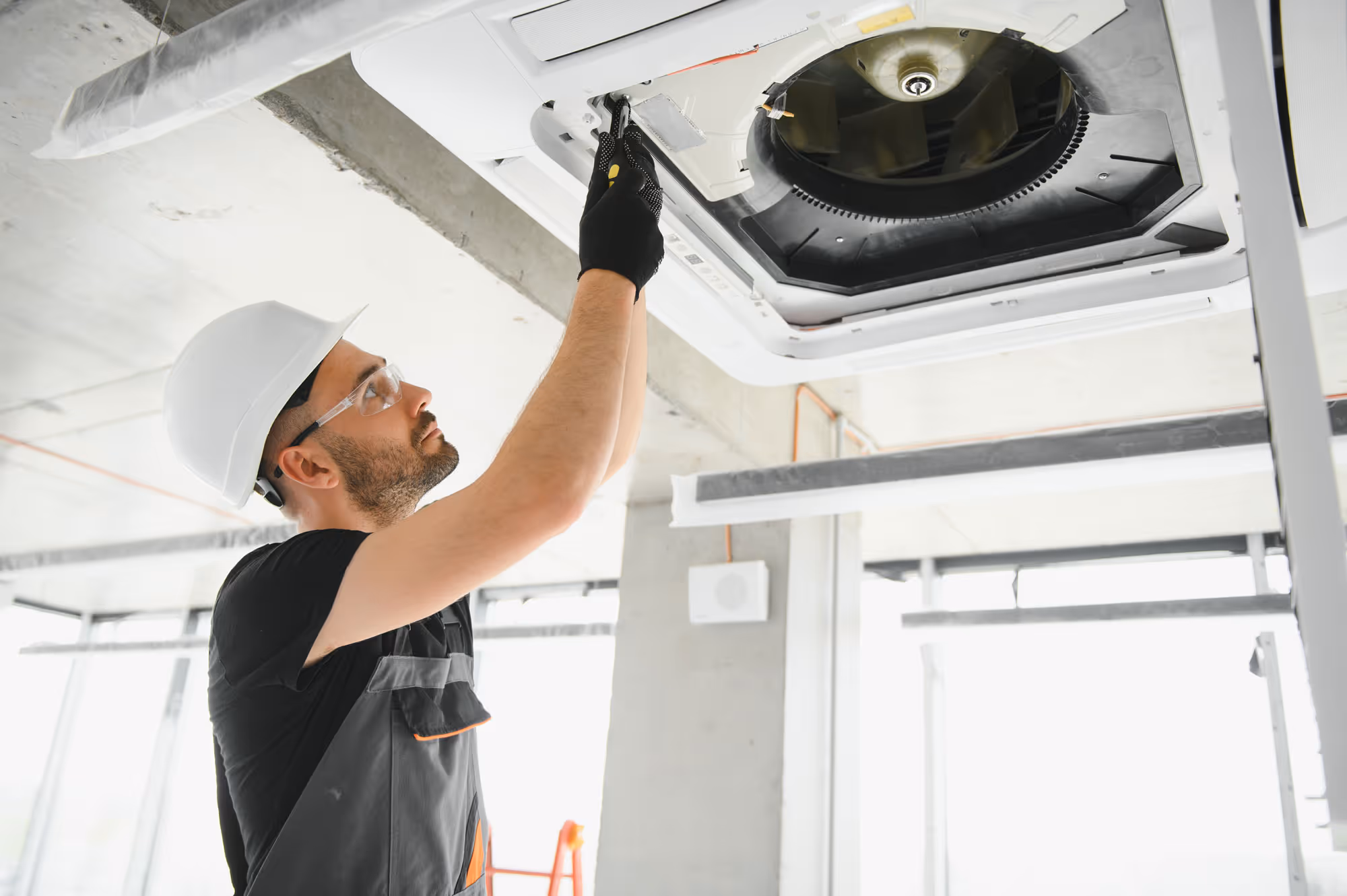 Un technicien portant un casque de protection et des lunettes de sécurité installe ou entretient un système de climatisation suspendu au plafond dans un bâtiment commercial en construction.