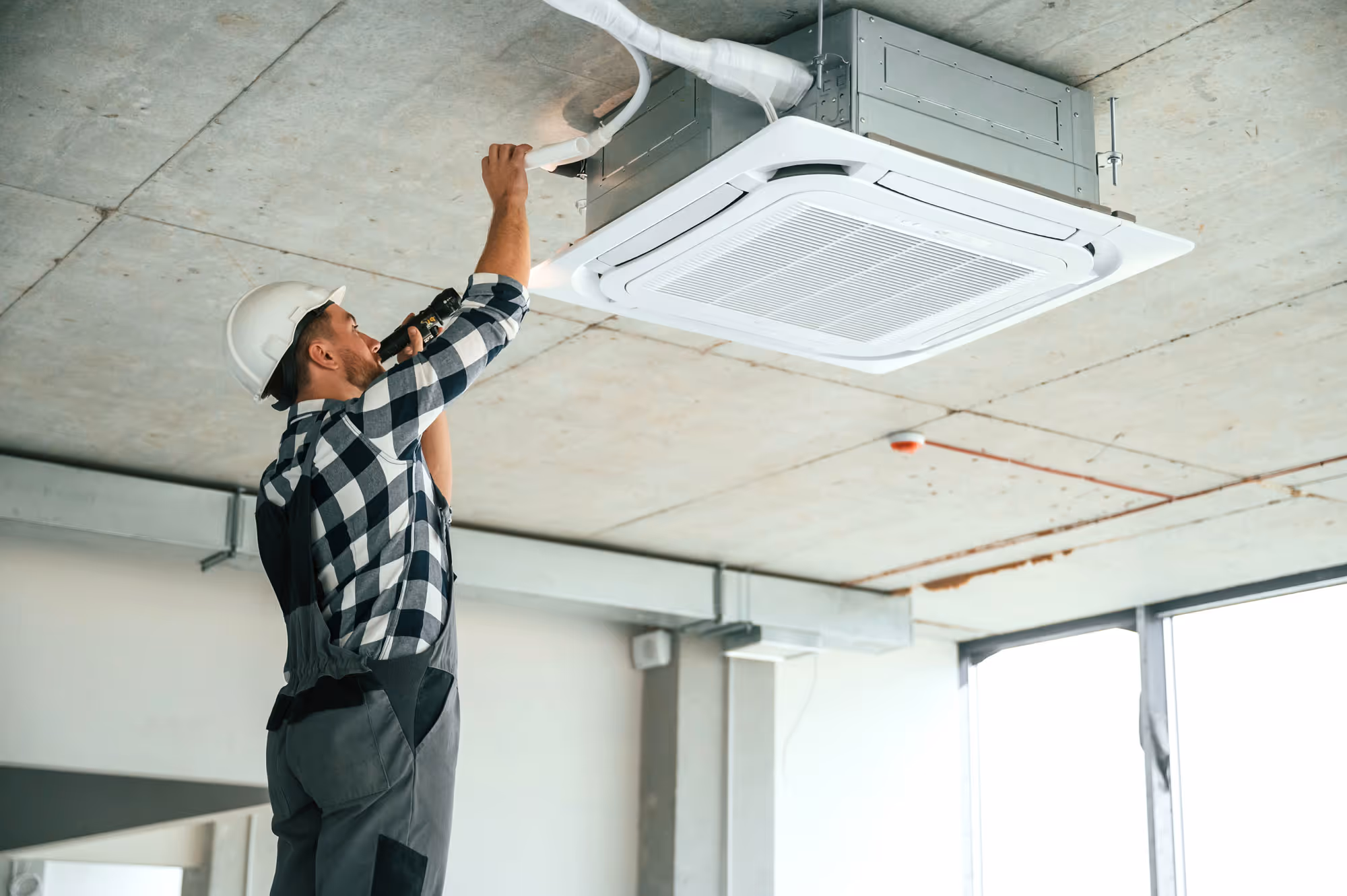 Technicien en casque blanc installant une unité de climatisation encastrée au plafond dans un bâtiment moderne avec une fenêtre à droite.