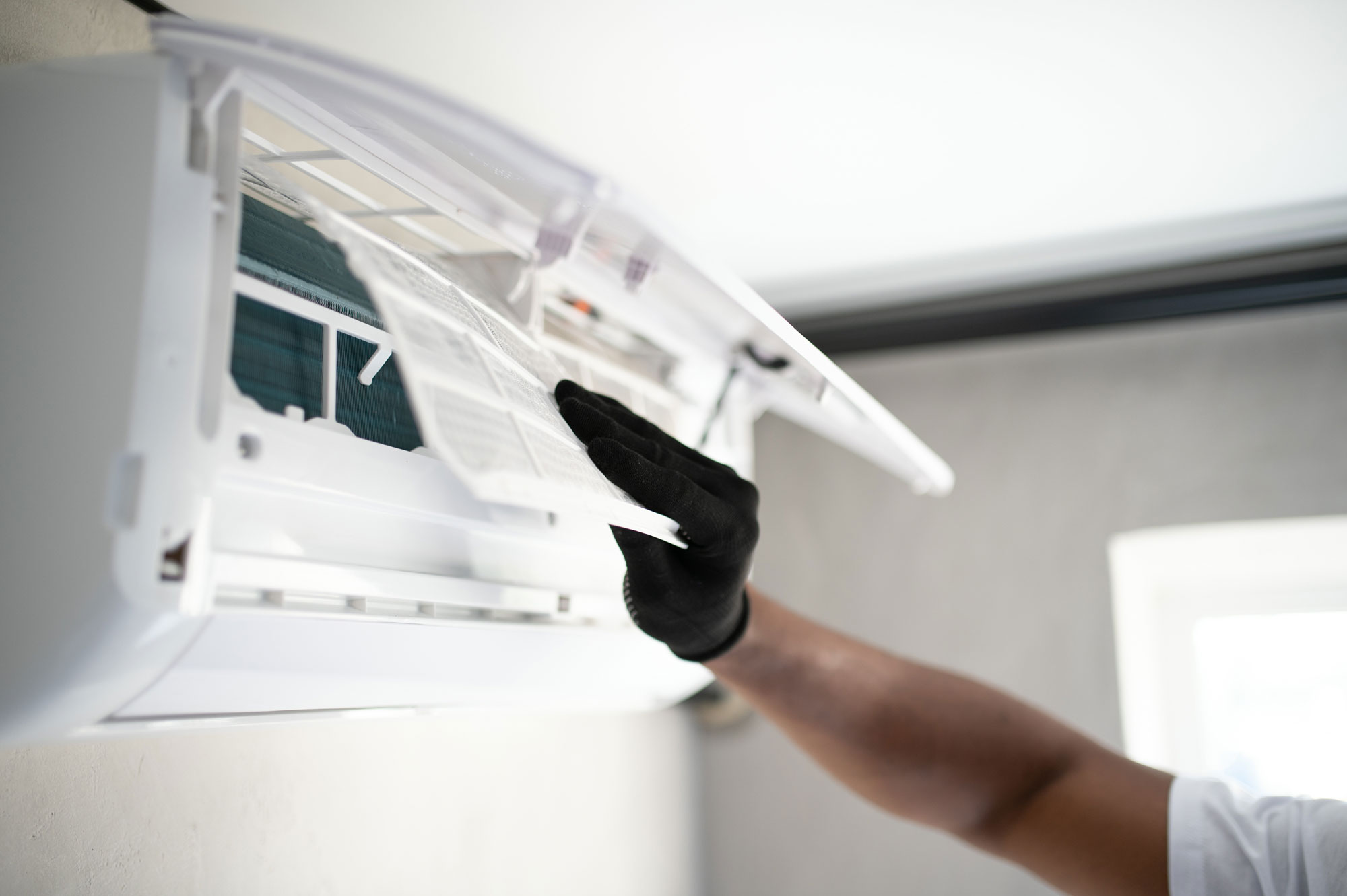 Une personne portant un gant noir nettoie ou inspecte le filtre d'un climatiseur mural blanc fixé au mur intérieur.