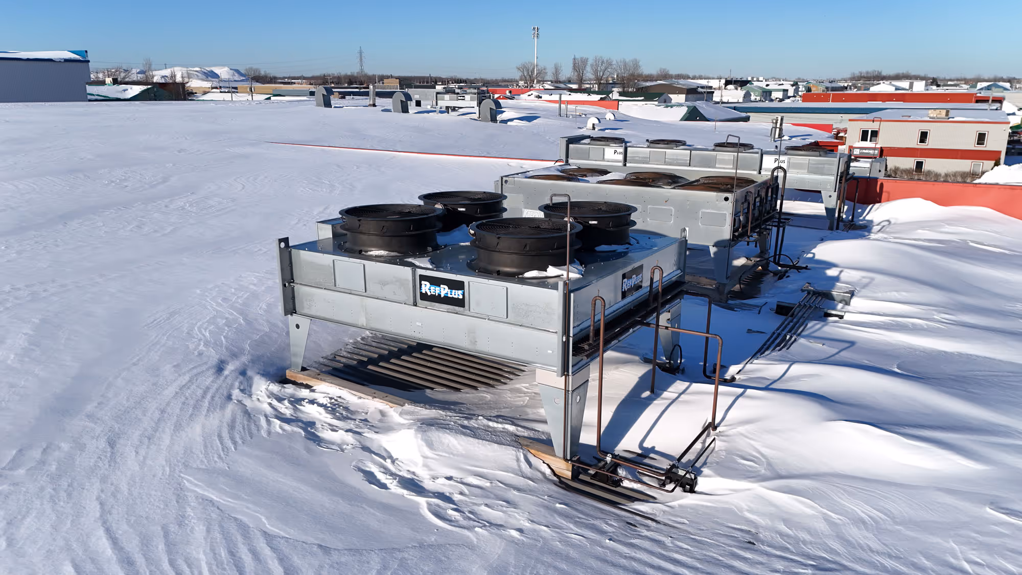 Unités de climatisation sur un toit plat recouvert de neige sous un ciel clair bleu, avec des bâtiments à l'horizon.