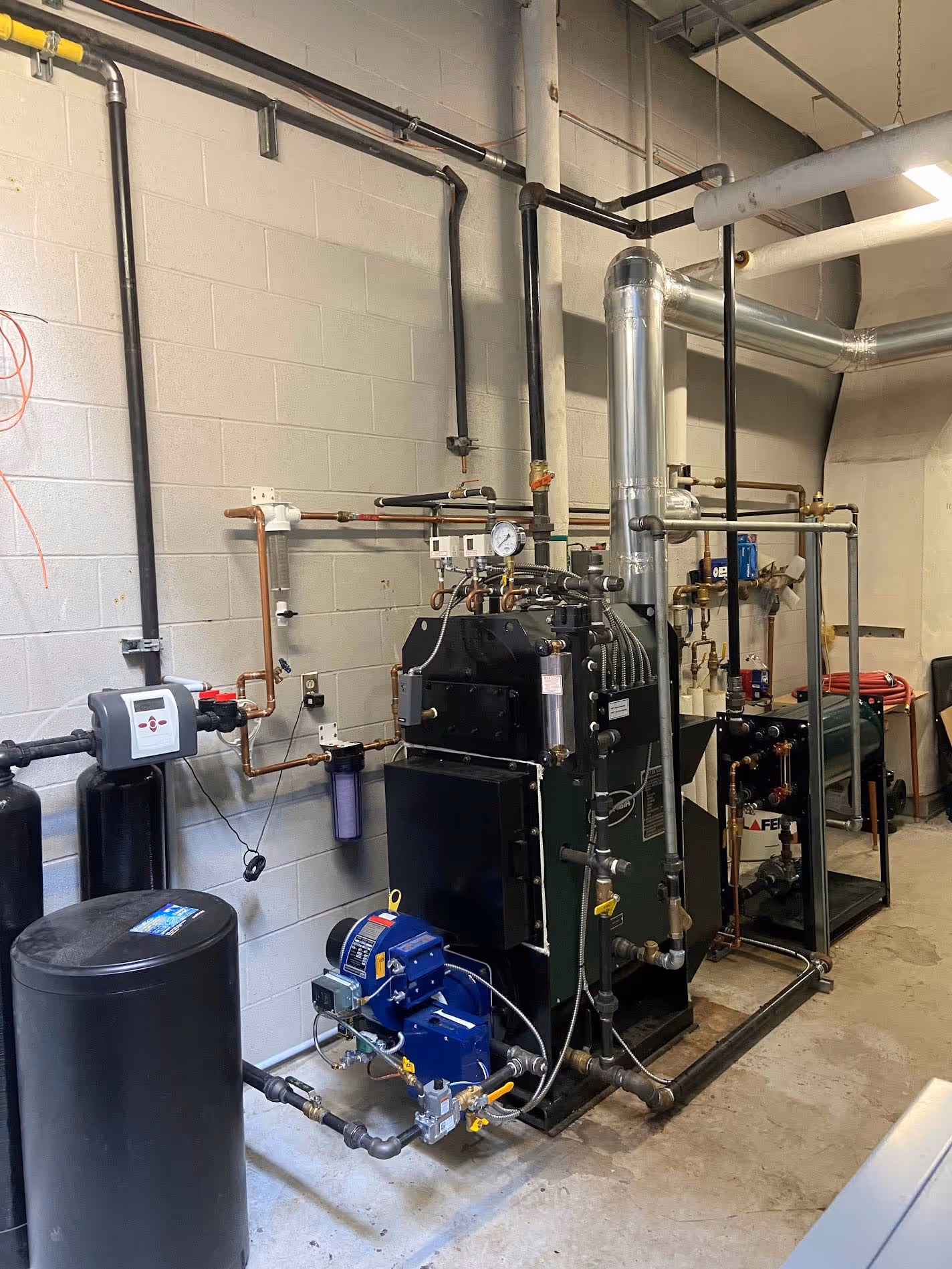Système de chaudière industriel avec tuyauterie en cuivre, système de filtration d'eau, unités de contrôle électrique et ventilateurs dans une salle technique au sol en béton.