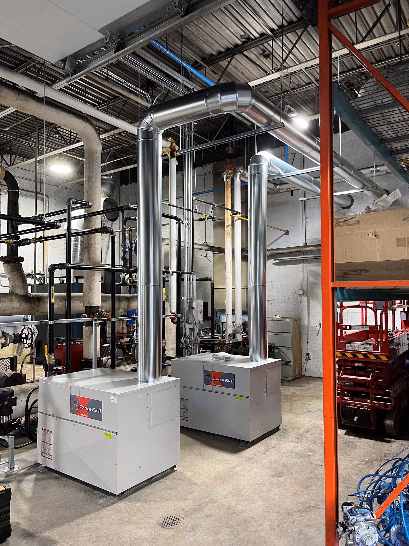 Salle industrielle avec deux grandes unités de chauffage blanc Connect-Pin II avec conduits d'évacuation métalliques s'élevant vers le plafond, entourées de tuyaux noirs et de structures métalliques, dans un espace aux murs en béton et au sol en béton.
